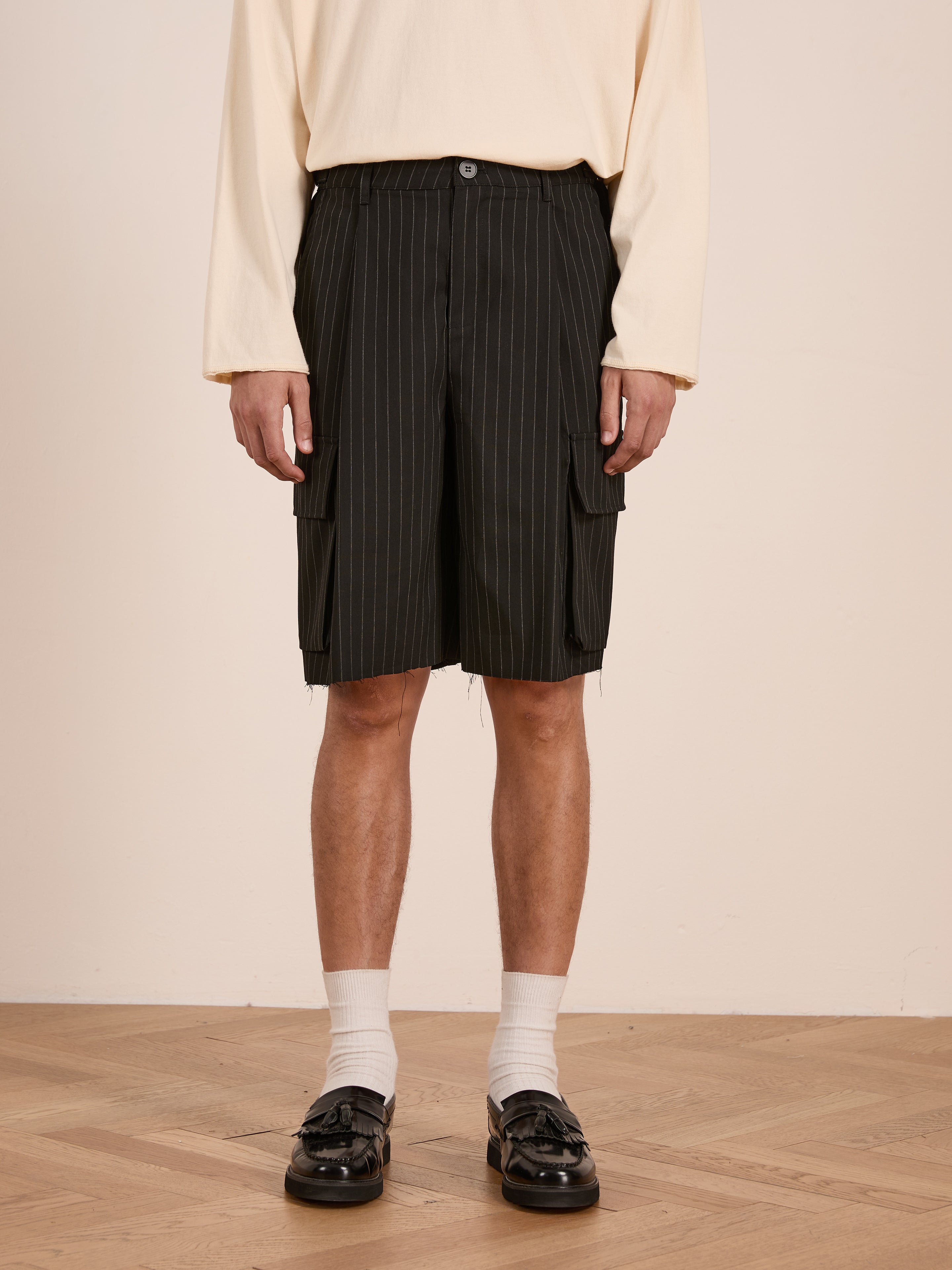 A person wears a cream long-sleeve shirt, FOUND Hava Pinstripe Pleated Cargo Trouser Shorts with an adjustable fit, white socks, and black loafers, standing on a wooden floor against a plain wall.
