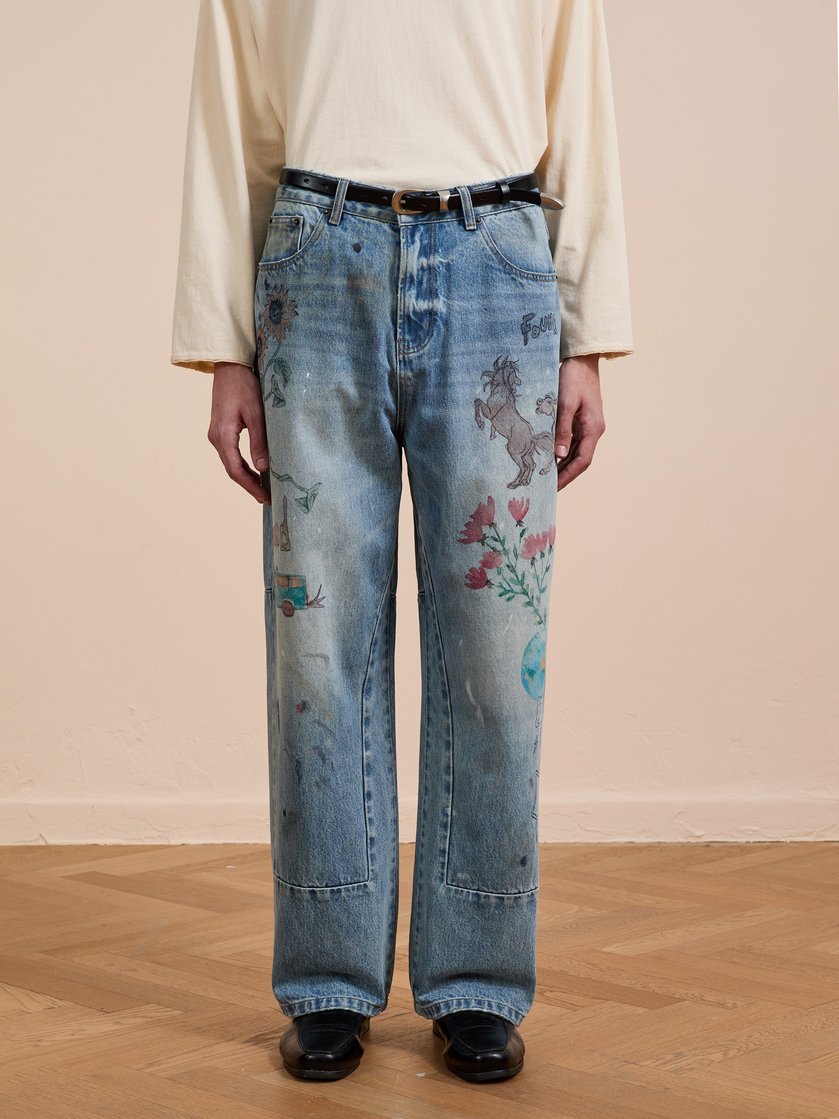 A person indoors wearing FOUND's Hand Art Jeans—loose denim with colorful, hand-drawn style prints—and a cream long-sleeve shirt; face not visible.