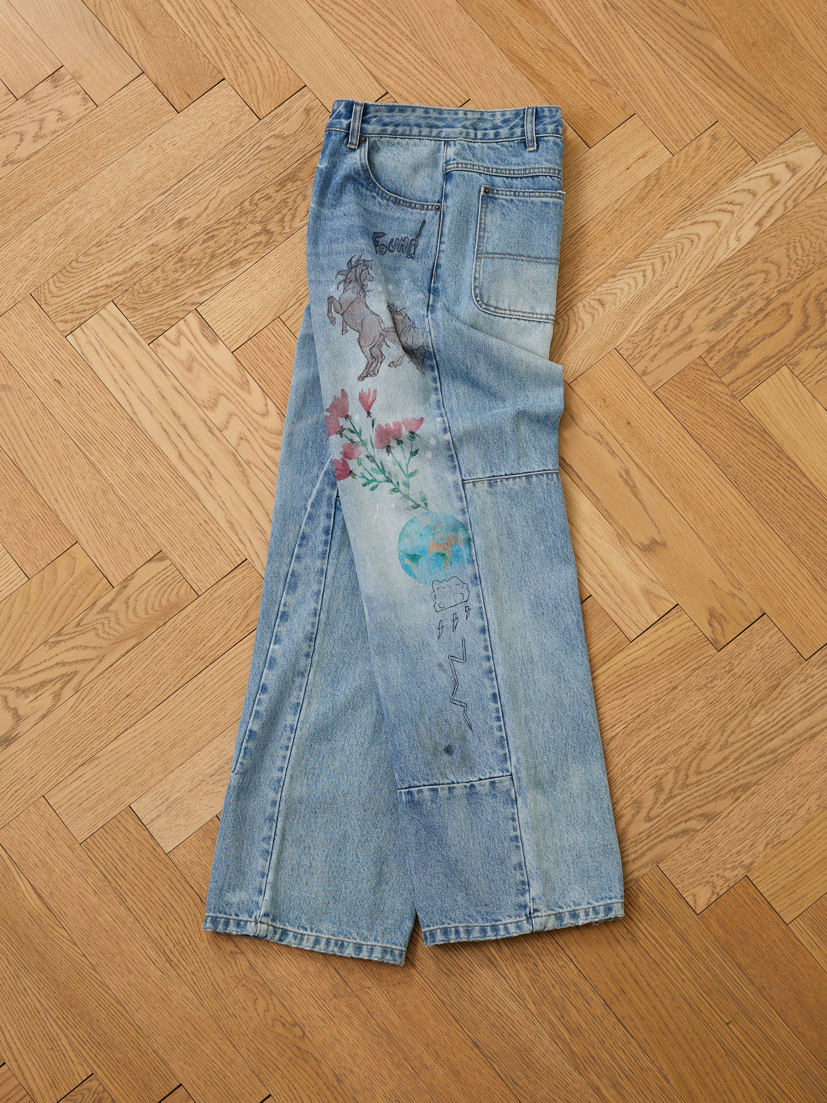 FOUND Hand Art Jeans in light blue denim, featuring original hand-drawn artwork of a horse, flowers, and the Earth, are shown laid flat on a wooden herringbone floor.