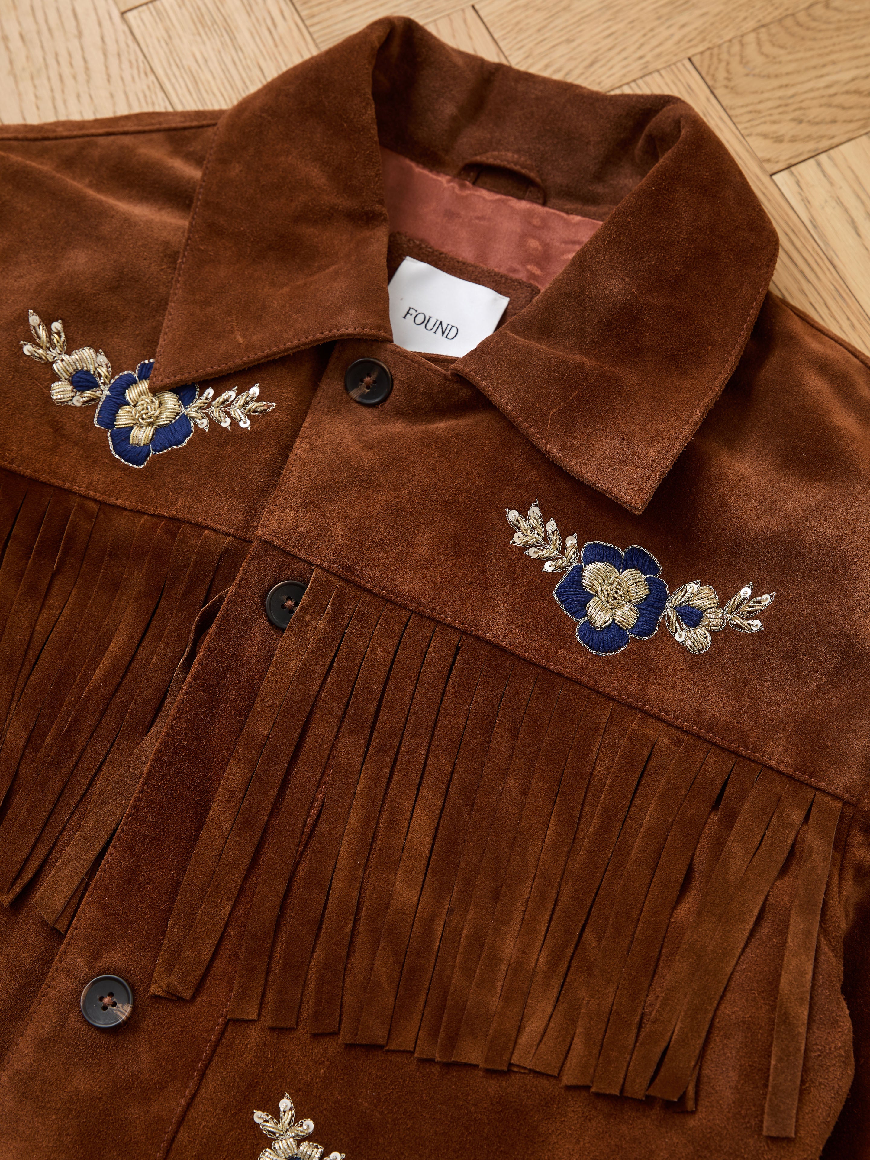 The FOUND Hand Embroidered Suede Fringe Jacket in brown features blue and gold floral embroidery on the chest and is displayed on a wooden floor.