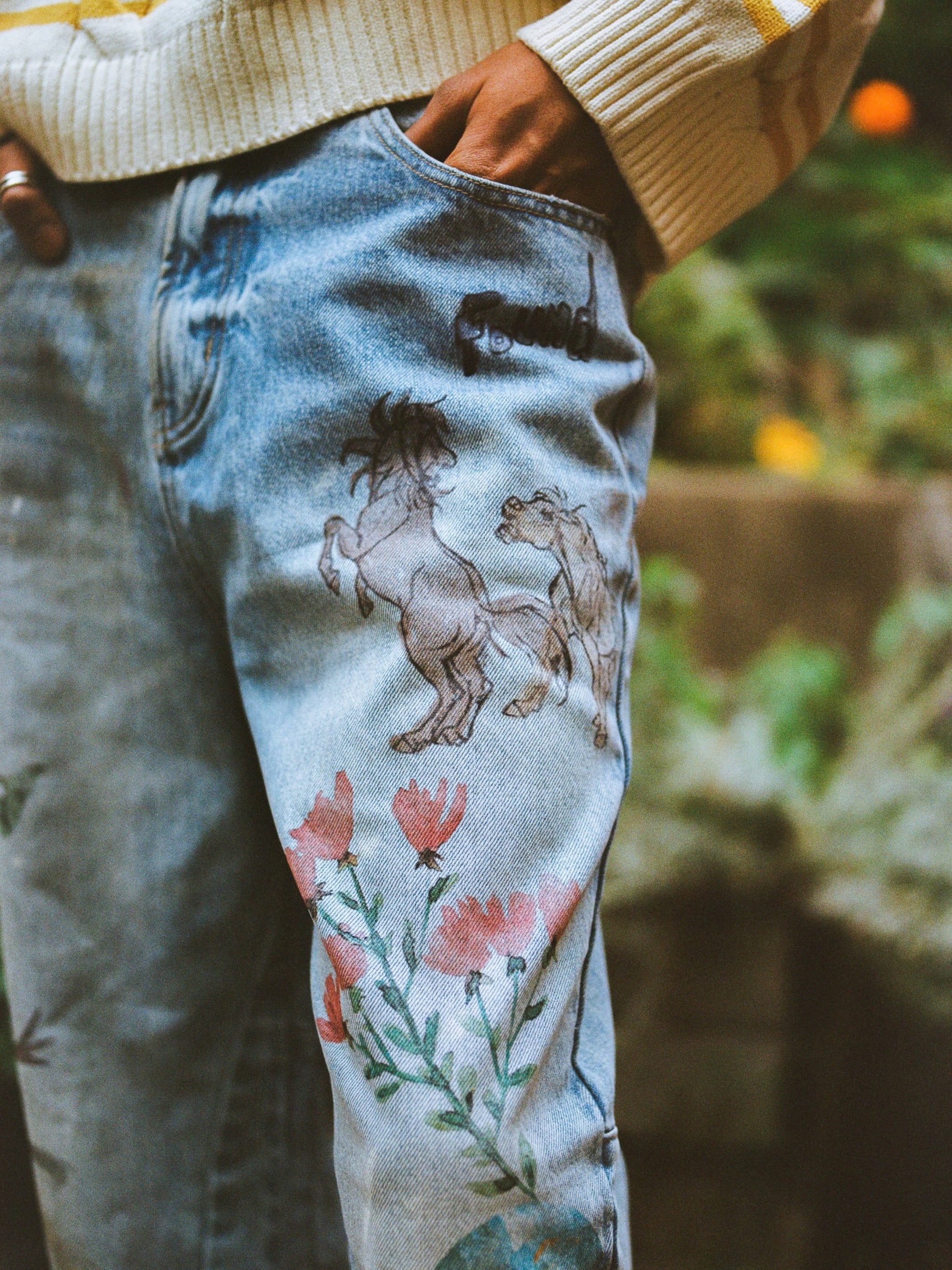 Close-up of a person wearing FOUND Hand Art Jeans in light blue denim with hand-drawn style horse and flower prints, paired with a cream sweater.