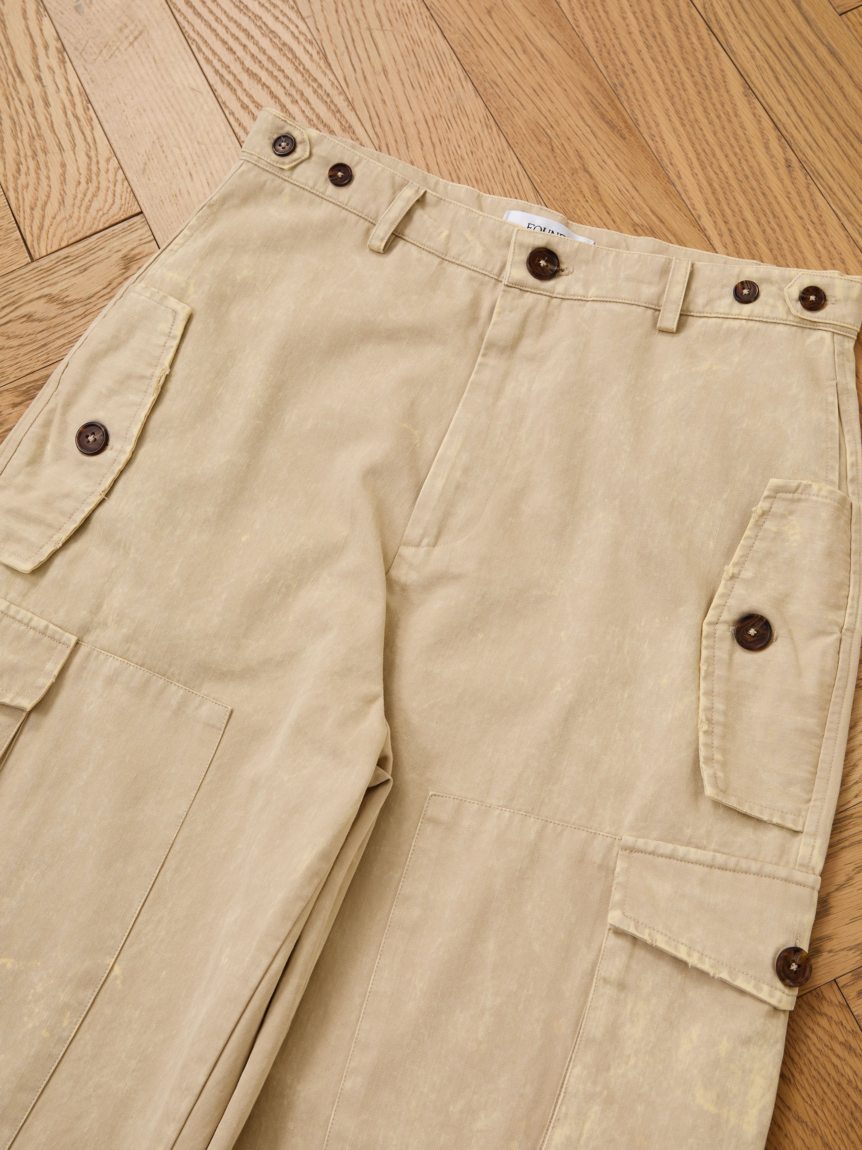 FOUND Utility Herringbone Paneled Pants in beige with side flap pockets, brown buttons, and a unisex relaxed fit, shown laid flat on a wooden herringbone floor.