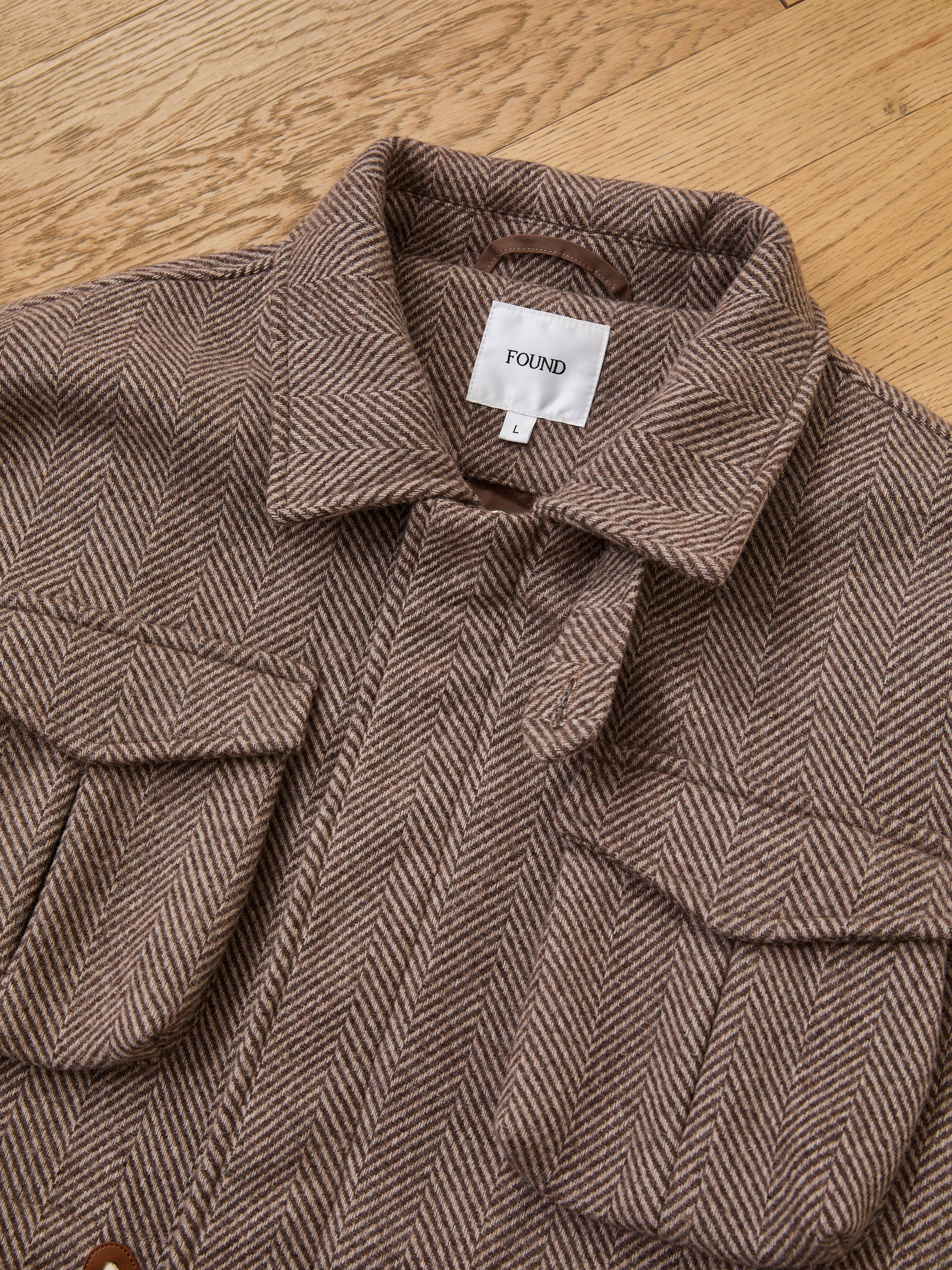 The FOUND Herringbone Wool Barn Jacket, made from a brown wool blend with two front pockets and a "FOUND" label on the collar, is displayed flat on a wooden surface.