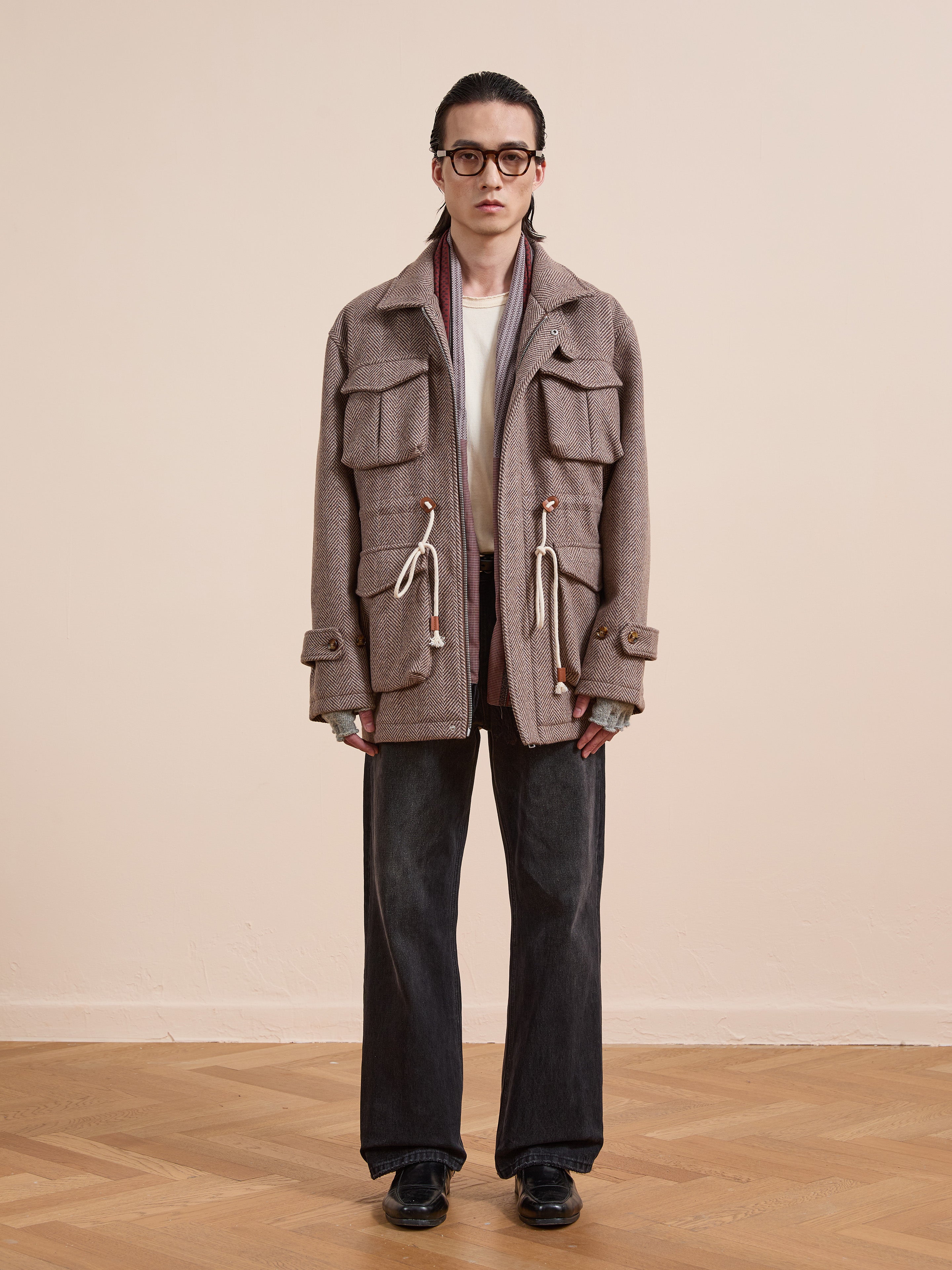 A person stands indoors against a plain background wearing glasses, a FOUND Herringbone Wool Barn Jacket in beige with large pockets and drawstrings, a white shirt, wide-leg dark jeans, and black shoes in a unisex relaxed fit style.