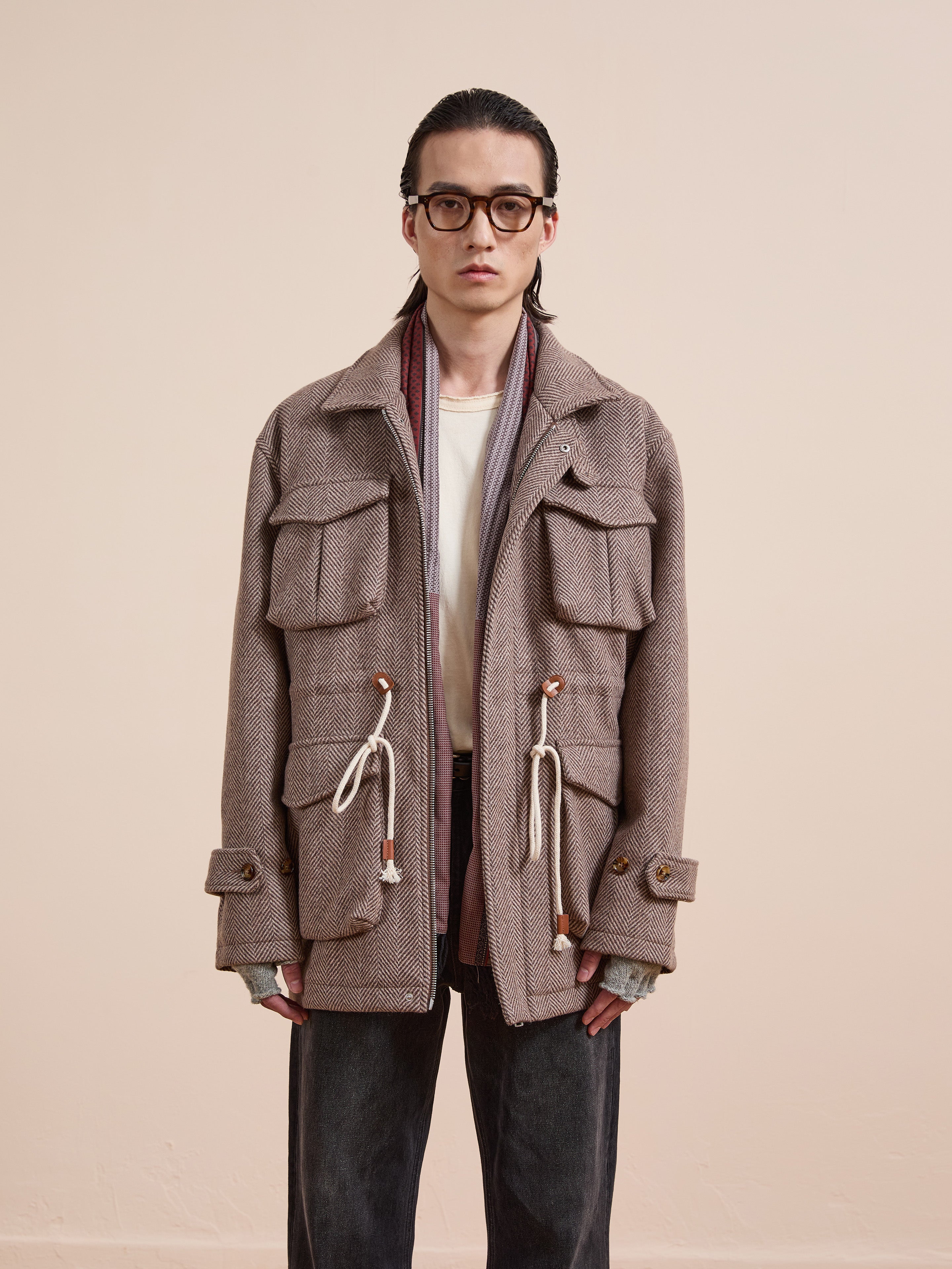 A person wears FOUND's Herringbone Wool Barn Jacket in a unisex relaxed fit with large pockets and drawstrings, paired with glasses, a white t-shirt, and dark jeans against a plain light-colored background.