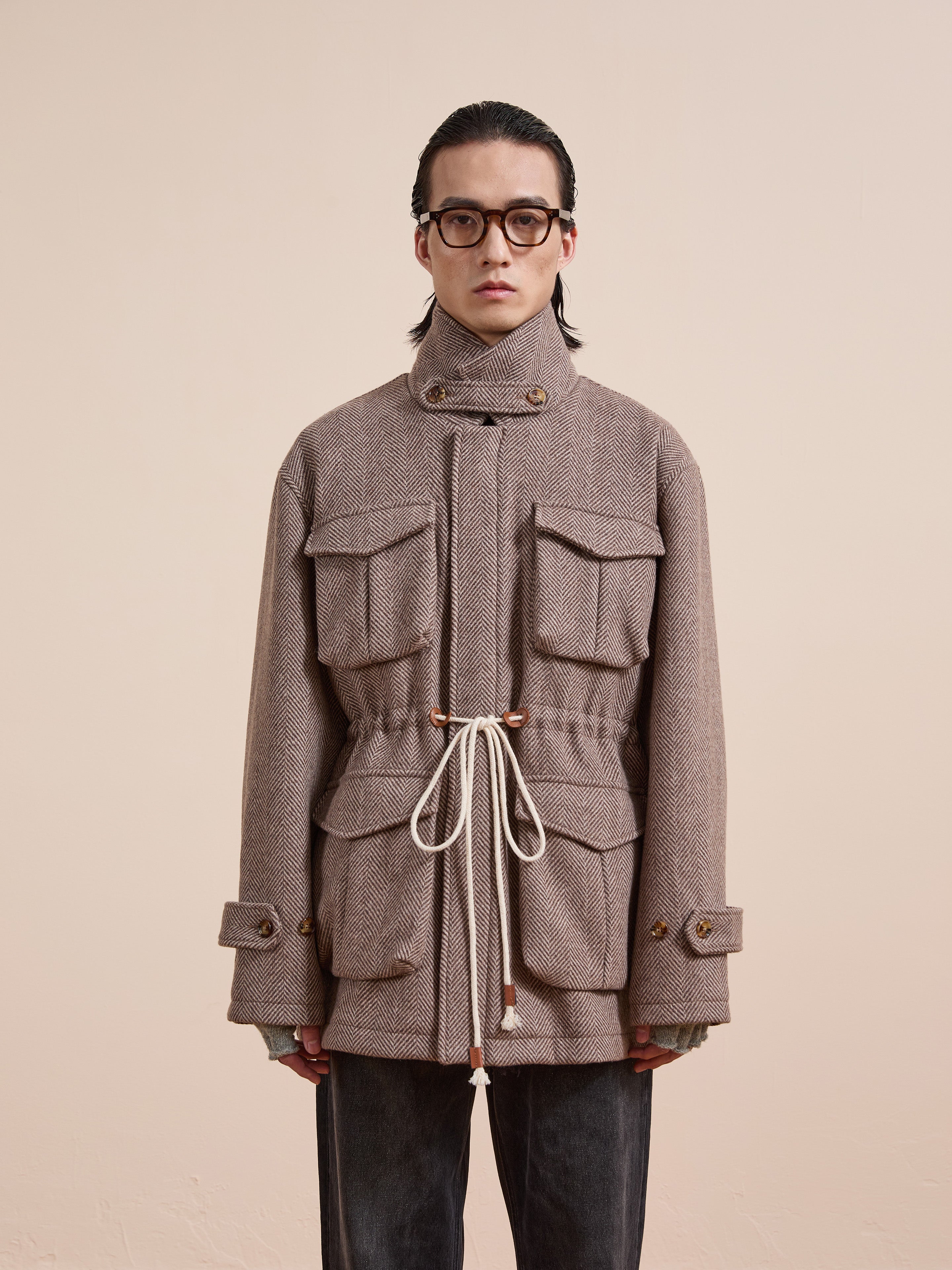 A person wearing glasses and the FOUND Herringbone Wool Barn Jacket—a unisex, relaxed-fit utility piece in textured beige with front pockets and a white drawstring—stands against a plain light background.