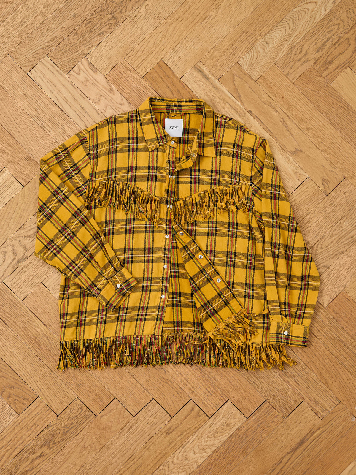 The High Plains Fringe Shirt by FOUND, a yellow plaid Western button-up with chest and hem fringe, is displayed flat on a light wood herringbone floor.