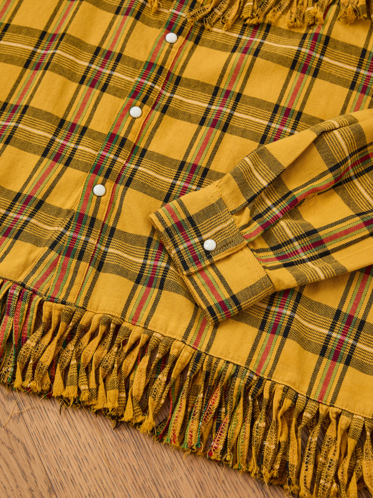 The FOUND High Plains Fringe Shirt, a yellow plaid unisex ranch style with white snaps and multicolored fringe hem, is shown on a wooden surface.