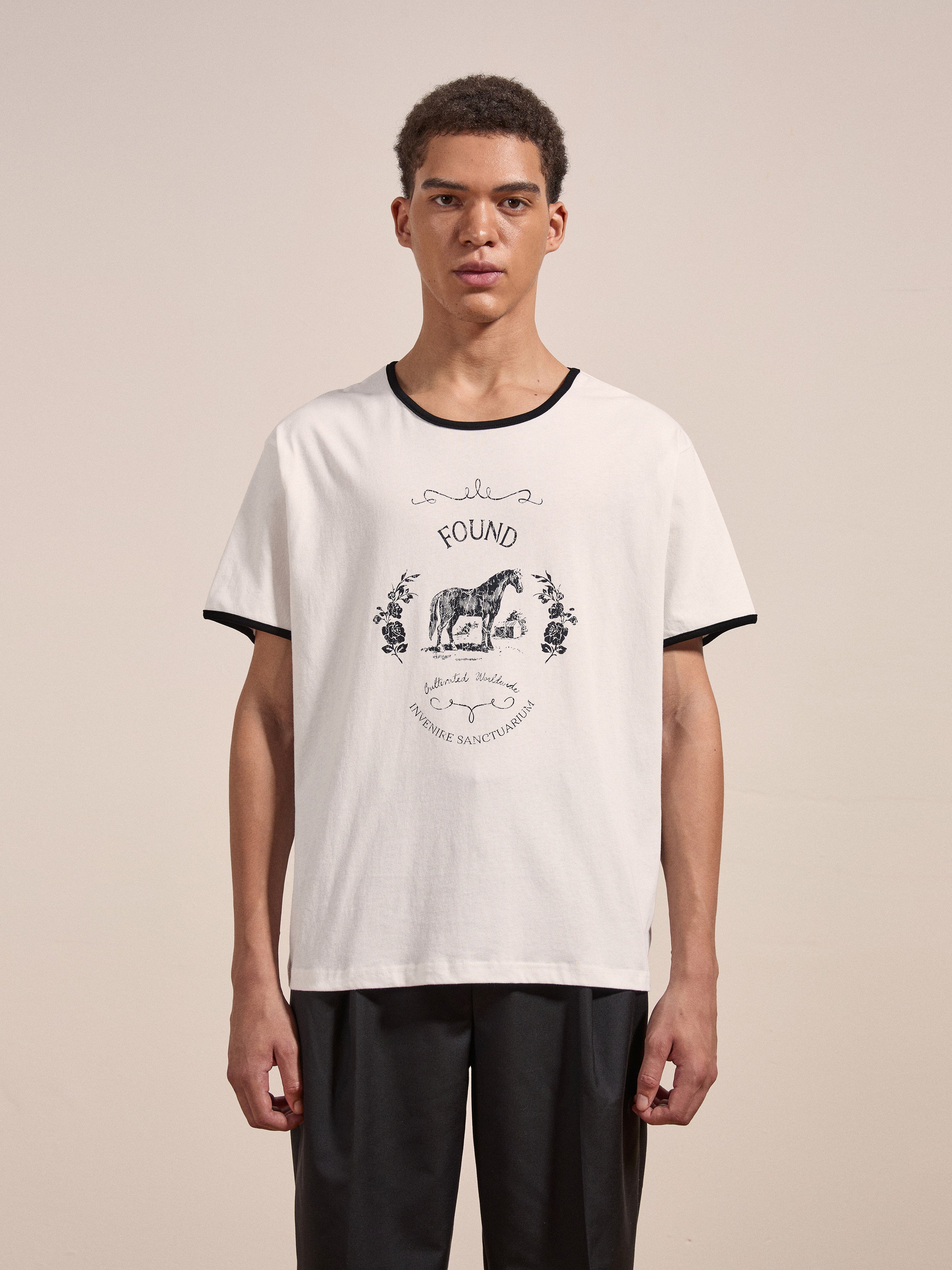 A young man wearing the FOUND Horse Crest Ringer Tee, featuring a white base with a black collar and an animal illustration on the front, stands against a plain background.