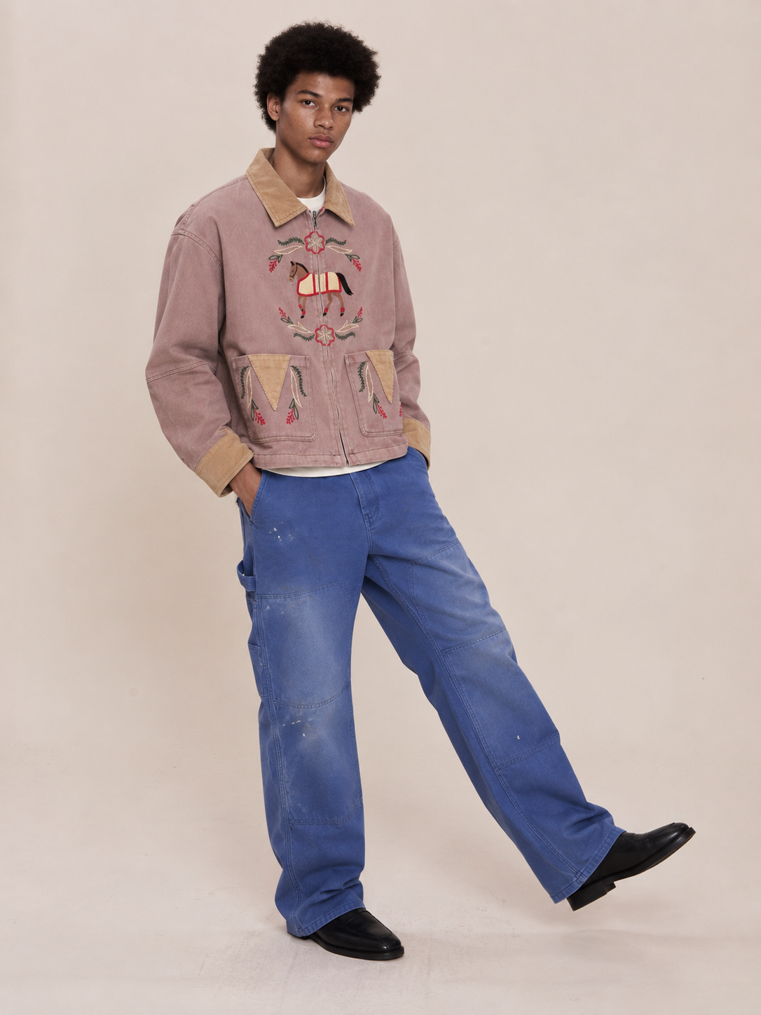 Person stands against a plain backdrop in the tan Horse Crest Work Jacket by FOUND with animal motifs, blue jeans, and black shoes—highlighting equestrian-inspired style.