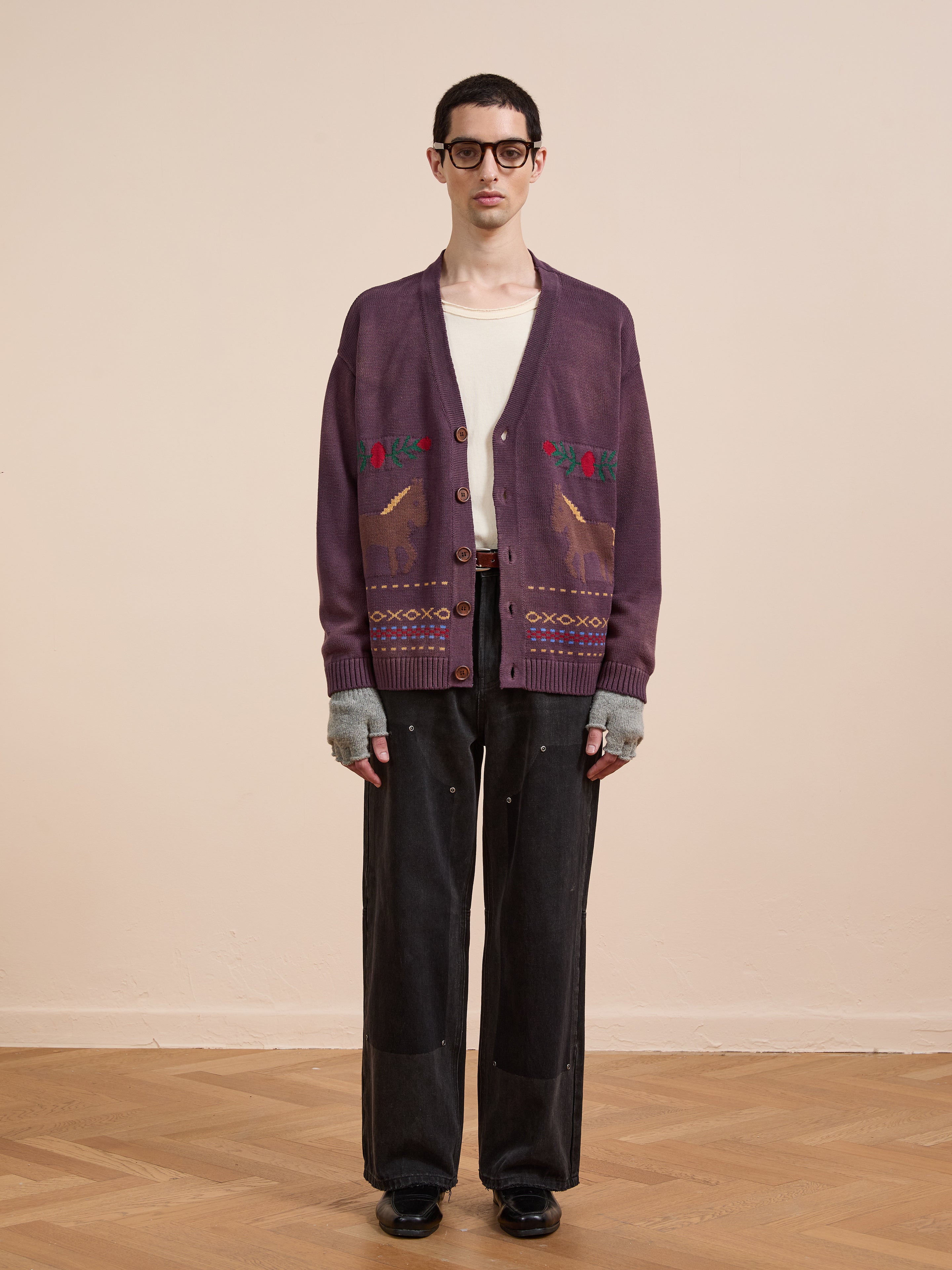 Indoors on a wooden floor, a person wears glasses, a Found Horse Jacquard Cardigan, white shirt, black pants, gray fingerless gloves, and black shoes.