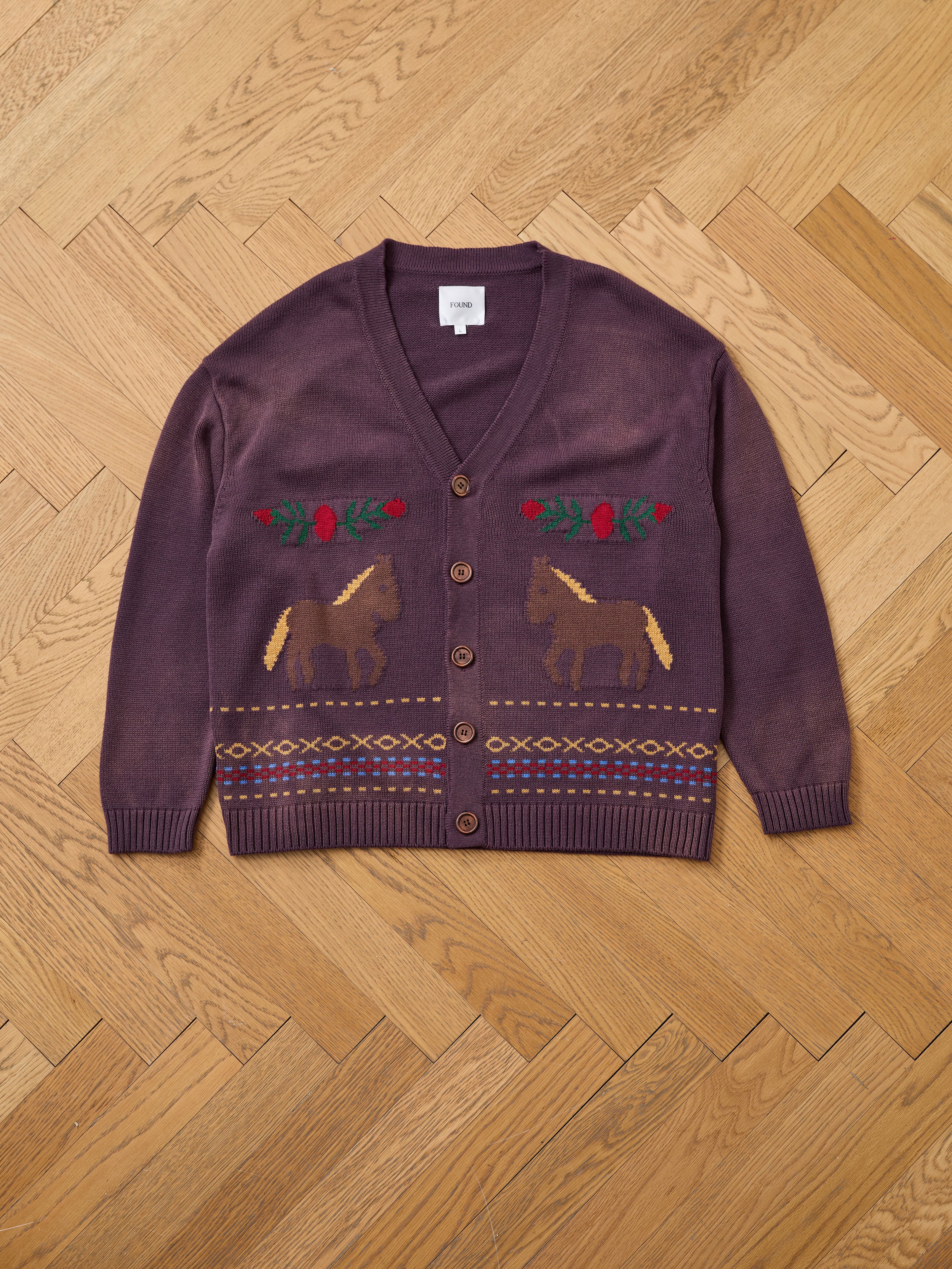 The Found Horse Jacquard Cardigan features a vintage purple design with a V-neck, brown buttons, red flowers, geometric patterns, and horse motifs, displayed flat on a wooden herringbone floor.