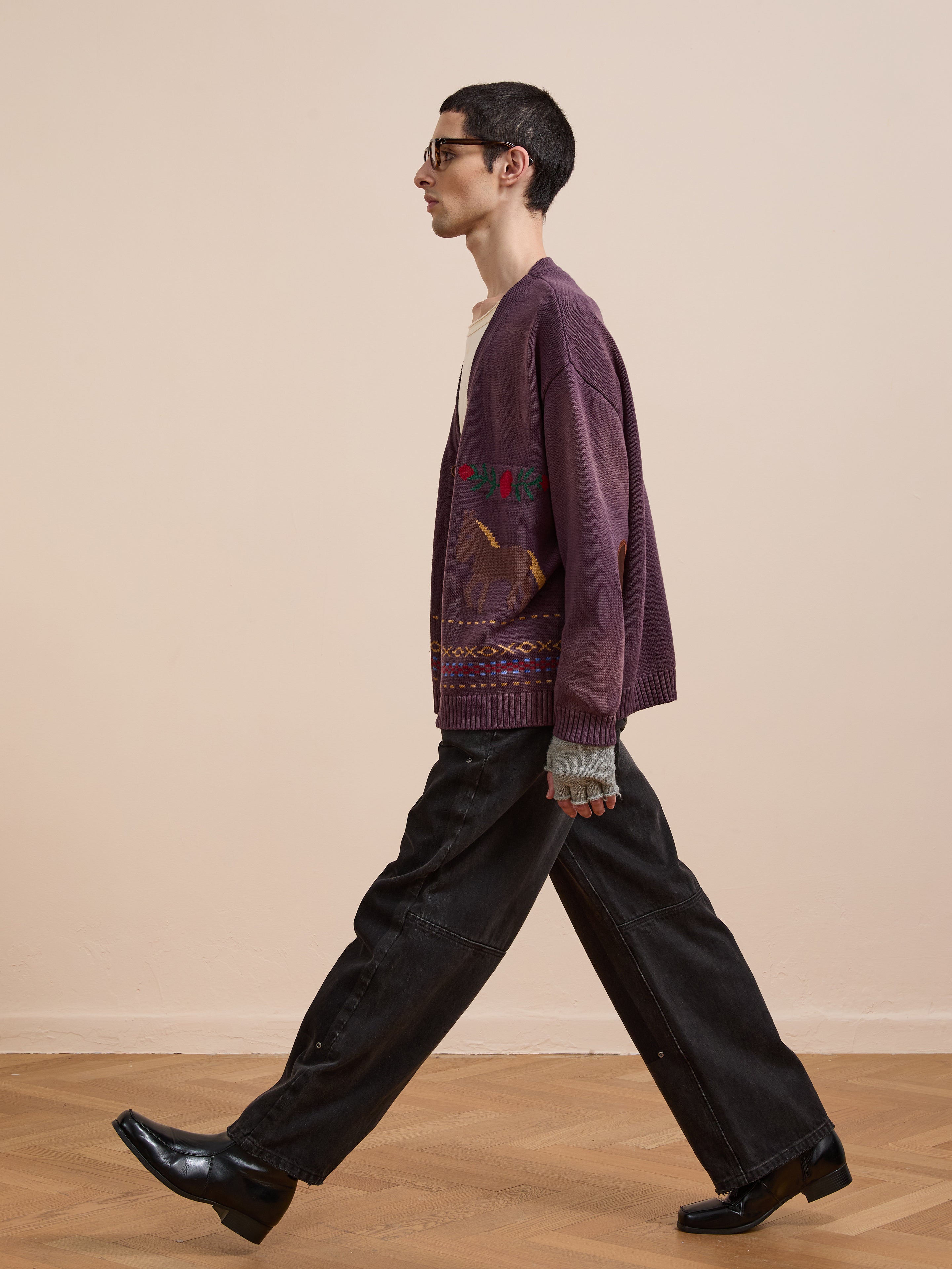 A person in profile walks indoors, wearing the Found Horse Jacquard Cardigan, loose black jeans, black square-toe shoes, and glasses against a plain beige wall.