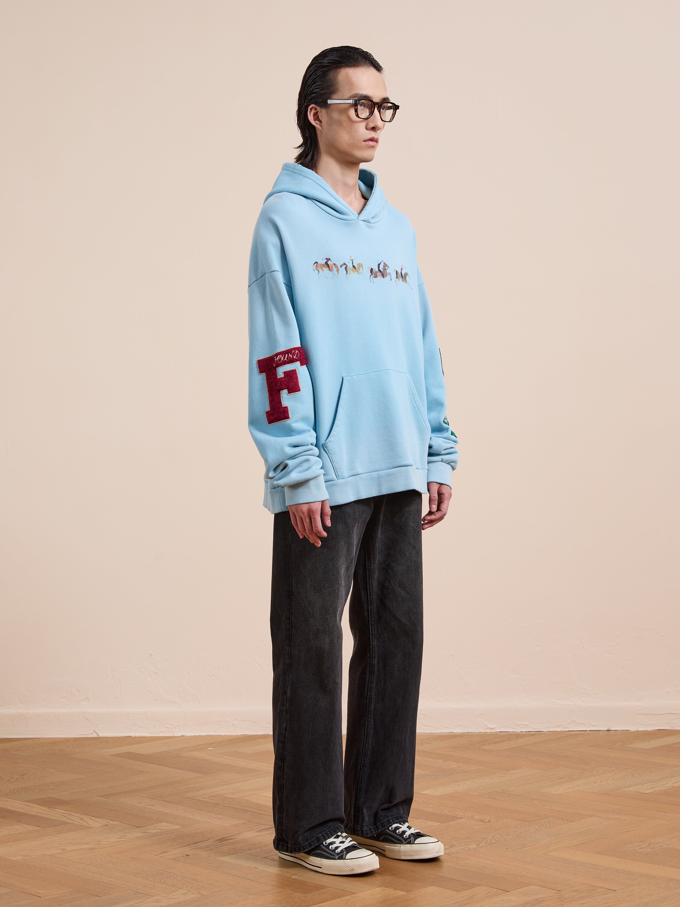 Indoors on a wooden floor, a person wears the FOUND Horse Line Hoodie in light blue French Terry cotton with embroidery, paired with black pants, black glasses, and black-and-white sneakers.