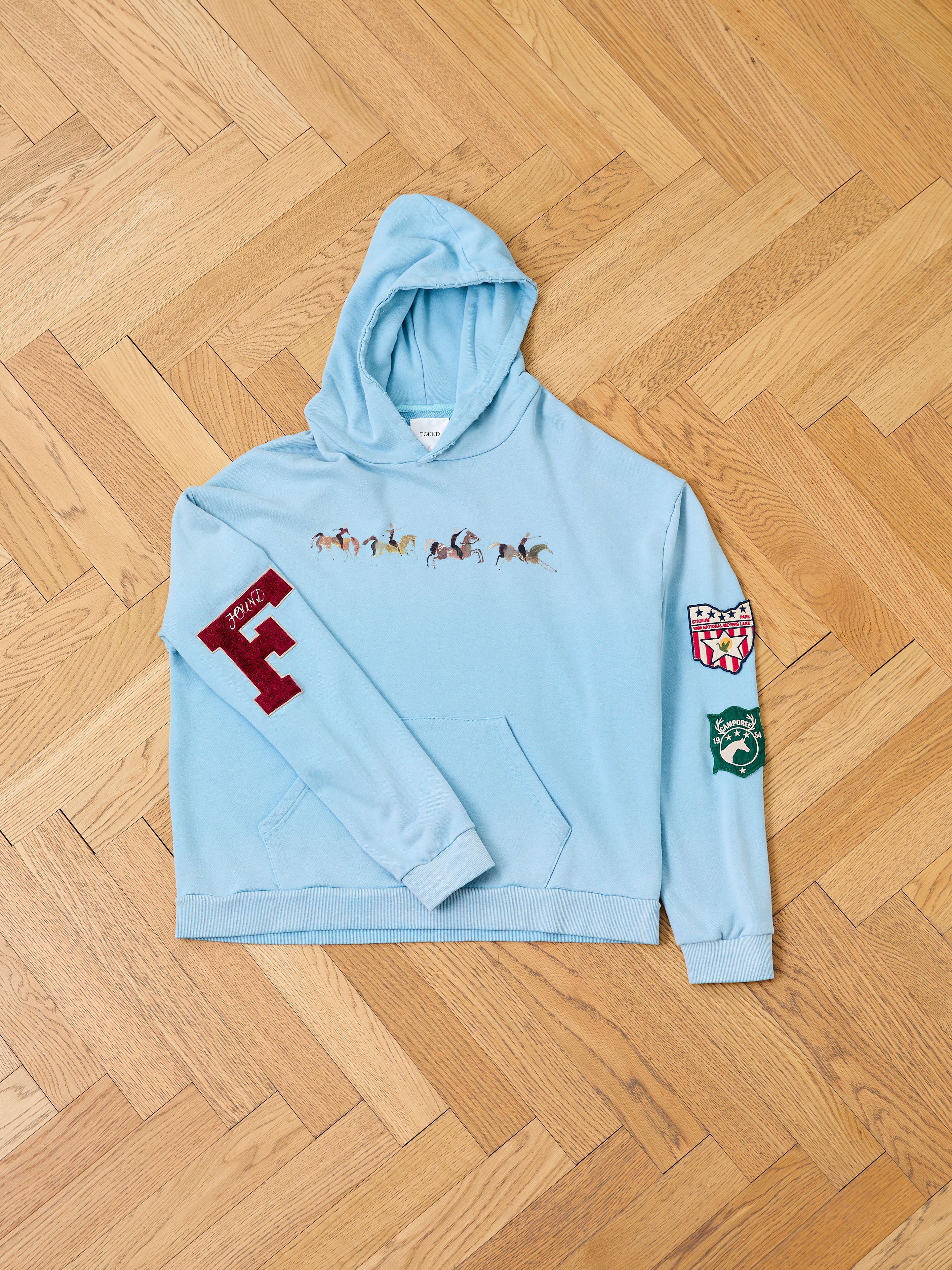 FOUND Horse Line Hoodie in light blue French Terry cotton, with small horse and rider chest illustrations, a large red "F" patch on one sleeve, and two extra patches on the other—shown on a wooden floor.