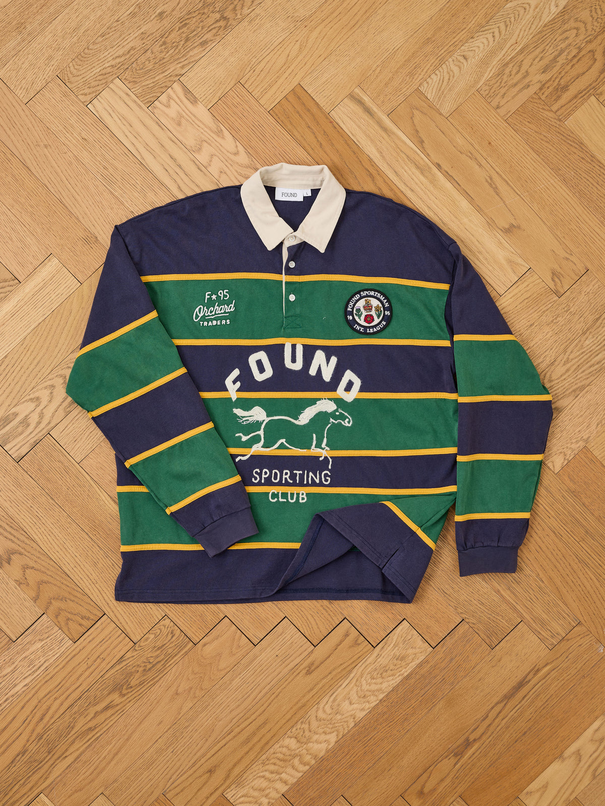 A unisex Horse Logo Rugby by FOUND featuring green, navy, and yellow stripes, white collar, and vintage team jersey style—laid flat on a wooden floor.