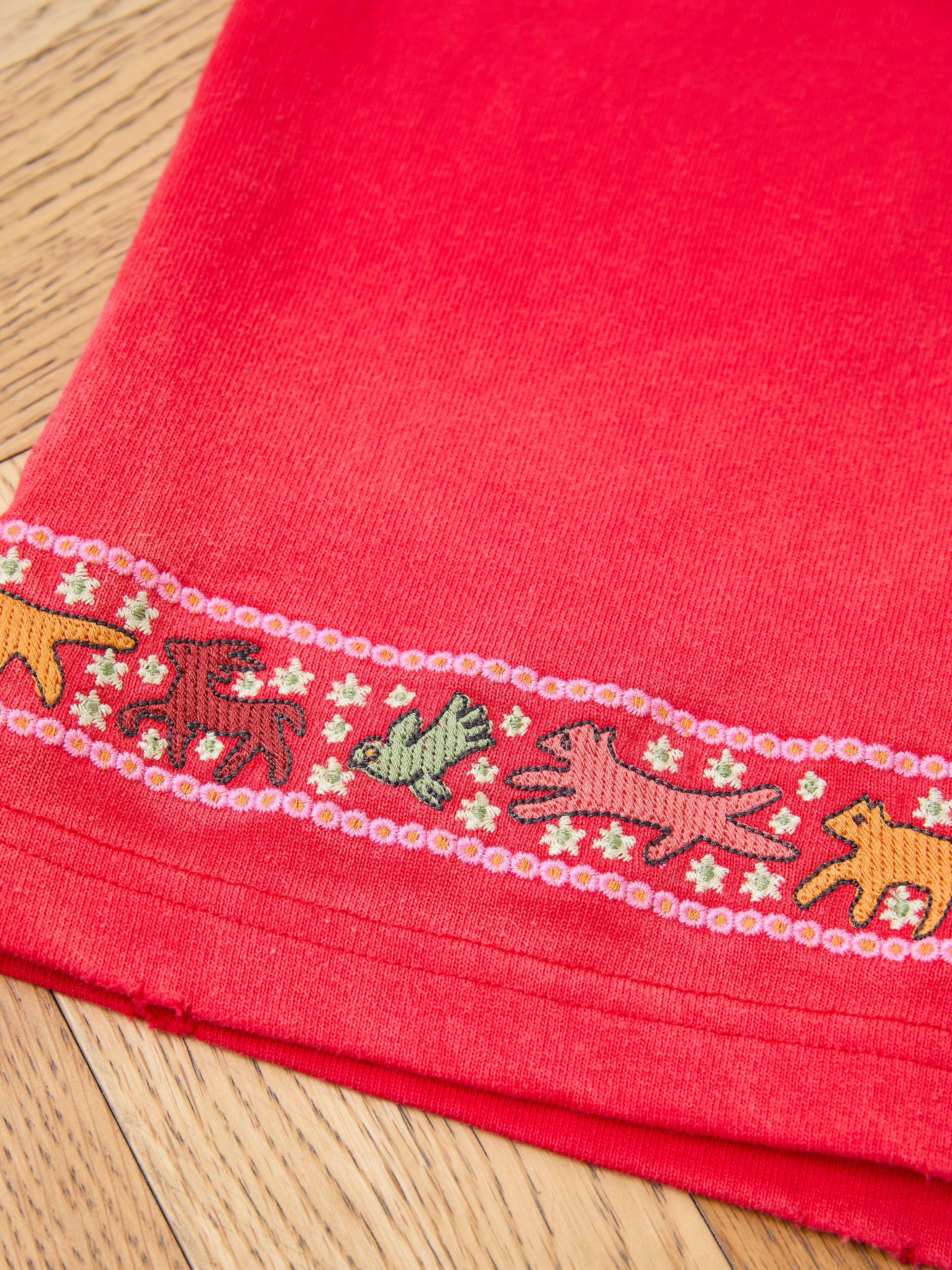 Close-up of the FOUND Horse Motif Embroidered Tee: red fabric with decorative trim and embroidered animals and flowers, laid on a wooden surface.