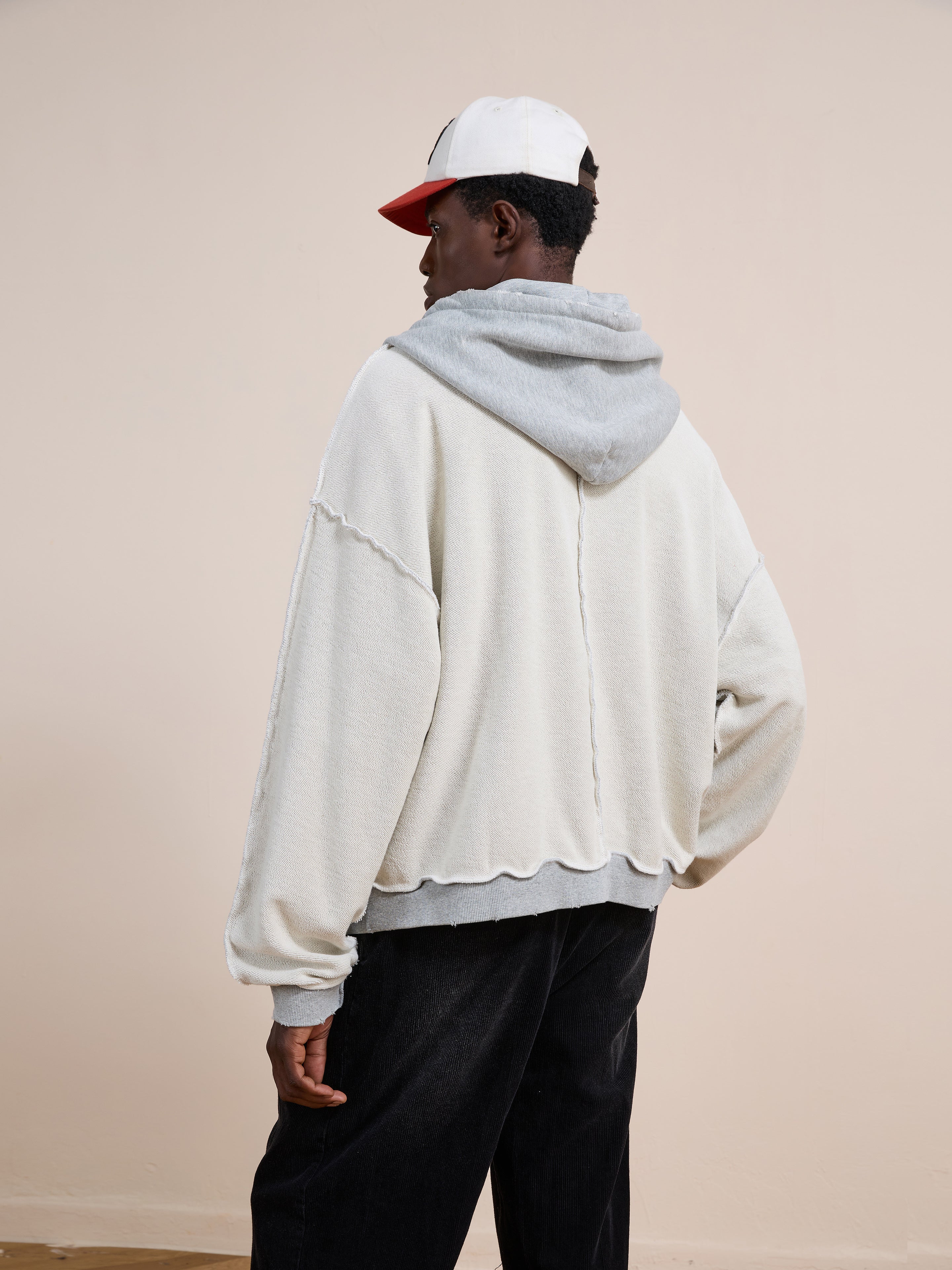 A person wearing the FOUND Deconstructed Terry Zip Up Hoodie with a gray hood stands with their back to the camera, paired with black pants and a white and red cap against a plain beige background.