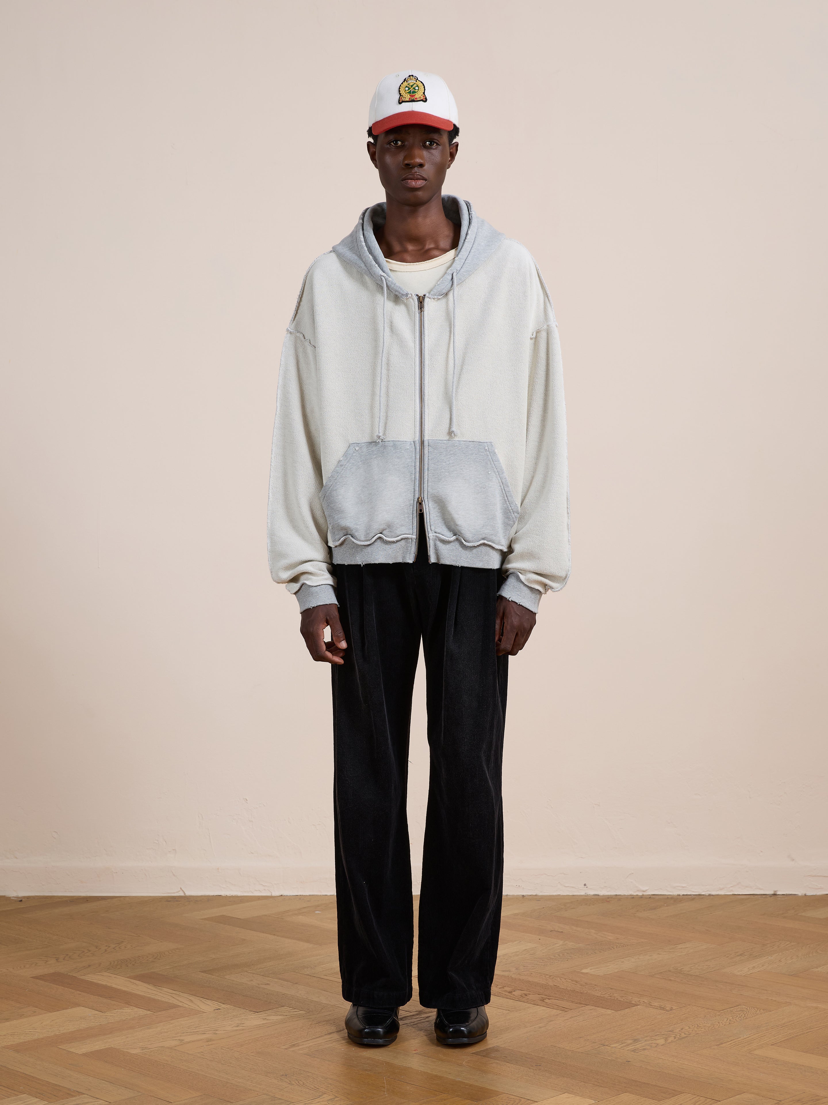 Indoors, a person wears the FOUND Deconstructed Terry Zip Up Hoodie in light gray, paired with wide-leg black pants, black shoes, and a white baseball cap with an embroidered patch.
