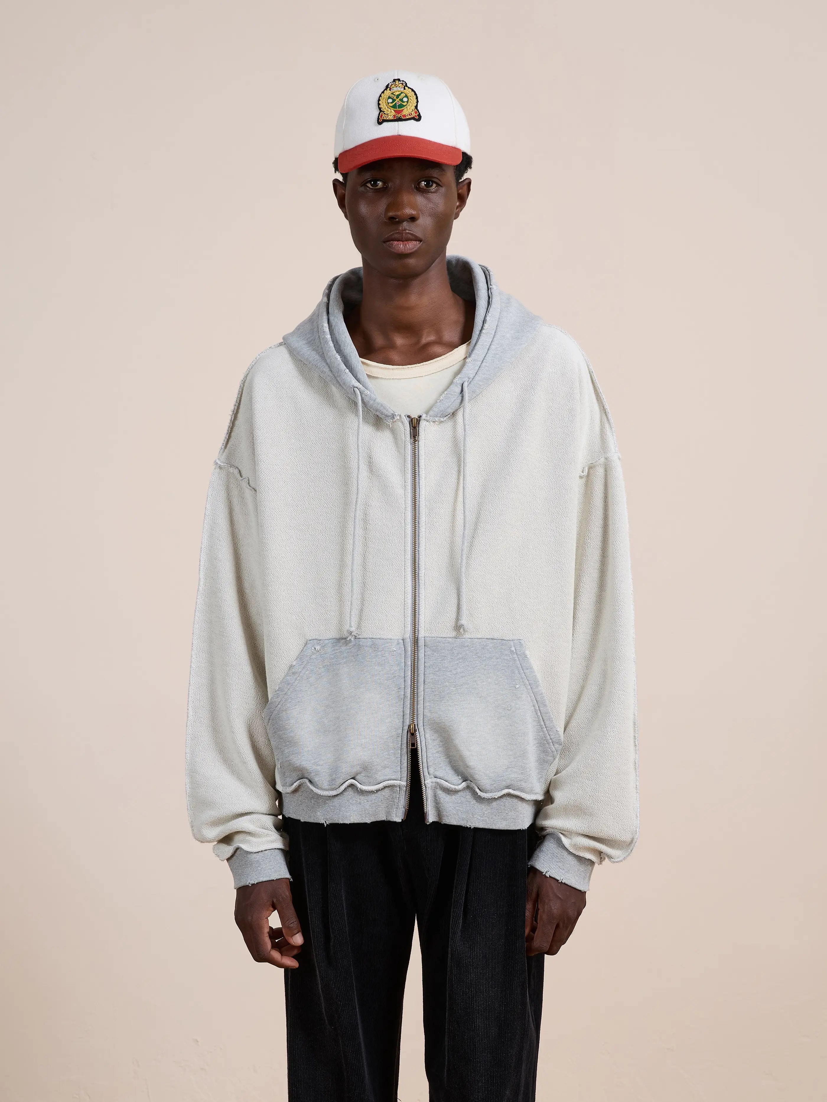 A person wears a white graphic baseball cap, a FOUND Deconstructed Terry Zip Up Hoodie in light grey, a white T-shirt, and black pants, standing against a plain beige background.