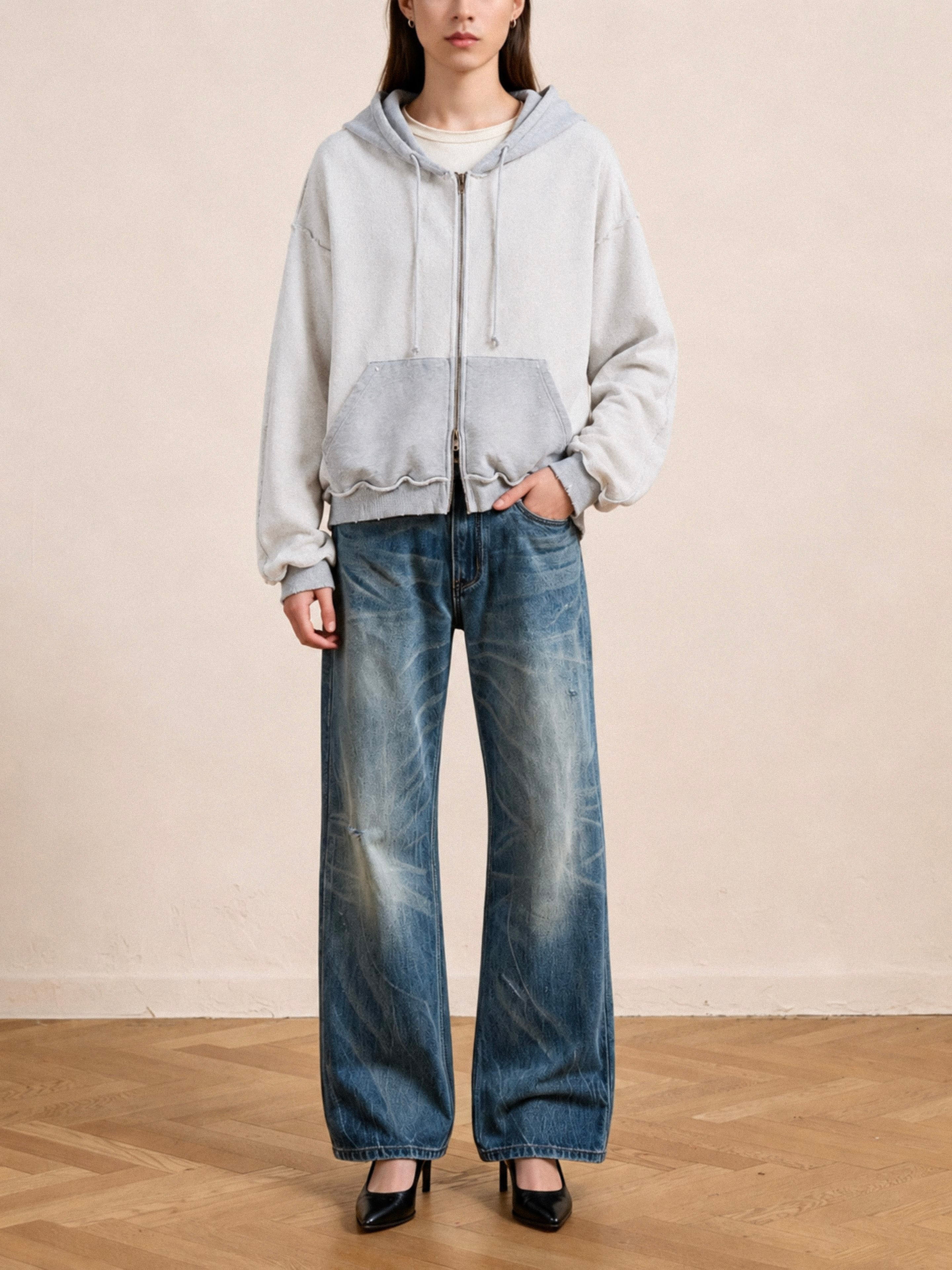 A person stands indoors on a wooden floor against a plain beige wall, wearing the FOUND Deconstructed Terry Zip Up Hoodie in light gray, loose blue jeans, and black heels.