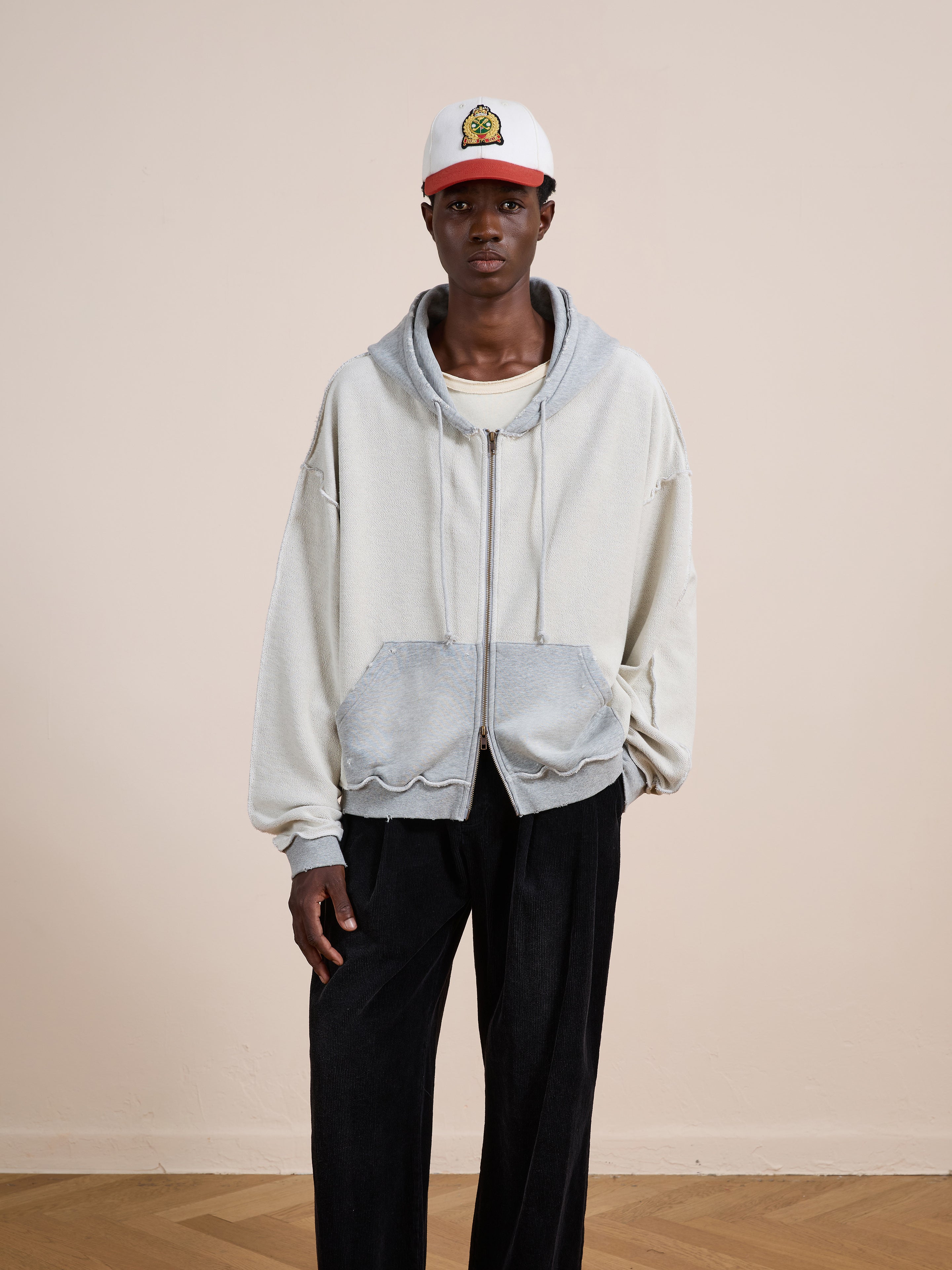 Indoors against a plain beige wall, a person wears the FOUND Deconstructed Terry Zip Up Hoodie over a cream shirt, paired with loose black pants and a white baseball cap.