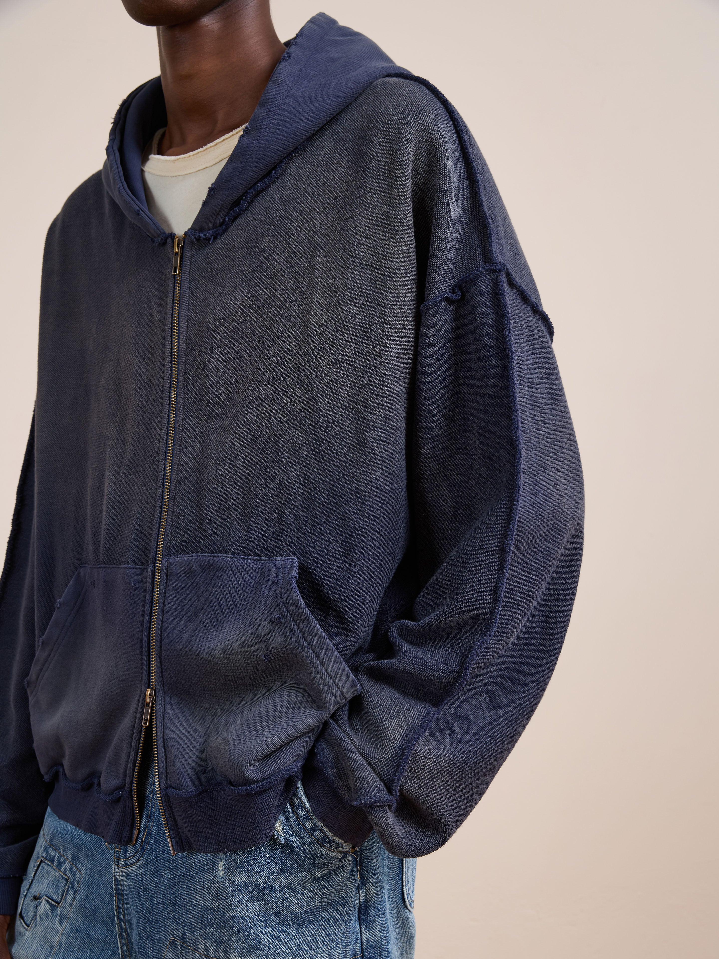 A person in a faded navy FOUND Deconstructed Terry Zip Up Hoodie over a beige t-shirt and blue jeans stands against a plain light background.