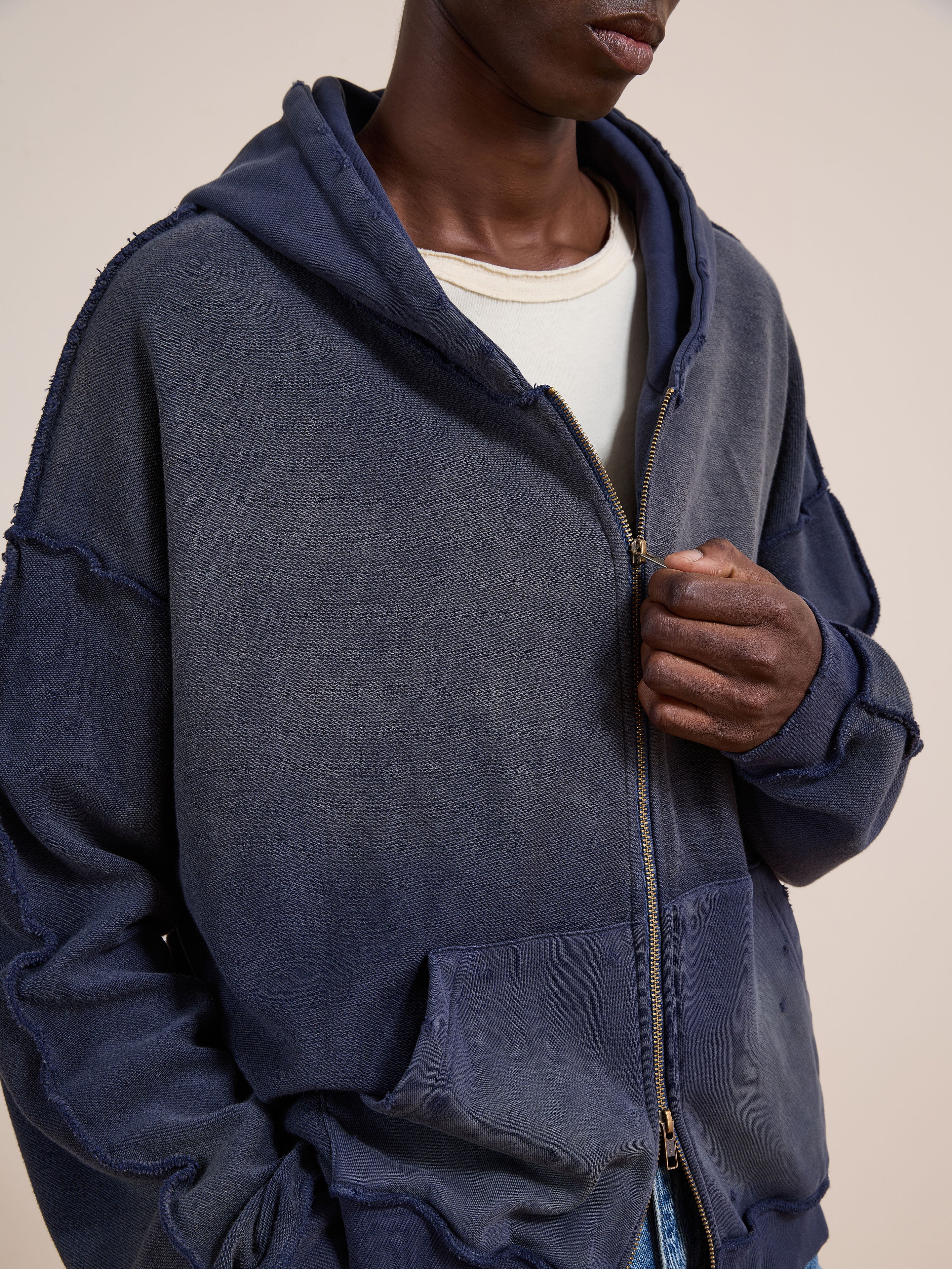 A person wears the FOUND Deconstructed Terry Zip Up Hoodie in faded navy blue over a white t-shirt, holding the zipper with one hand.