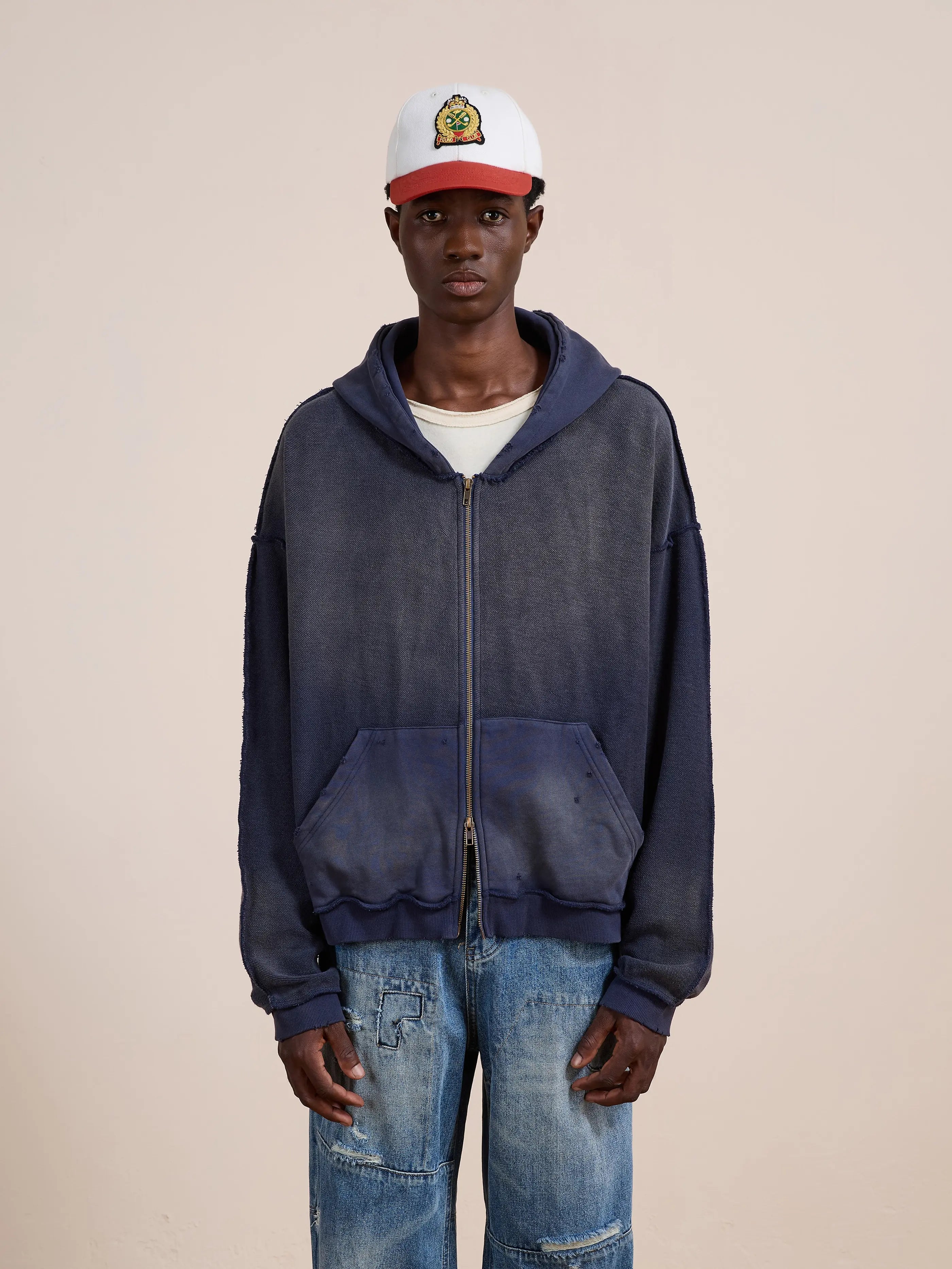 A person in a white and red cap, FOUND Deconstructed Terry Zip Up Hoodie, and distressed blue jeans stands against a plain beige background.