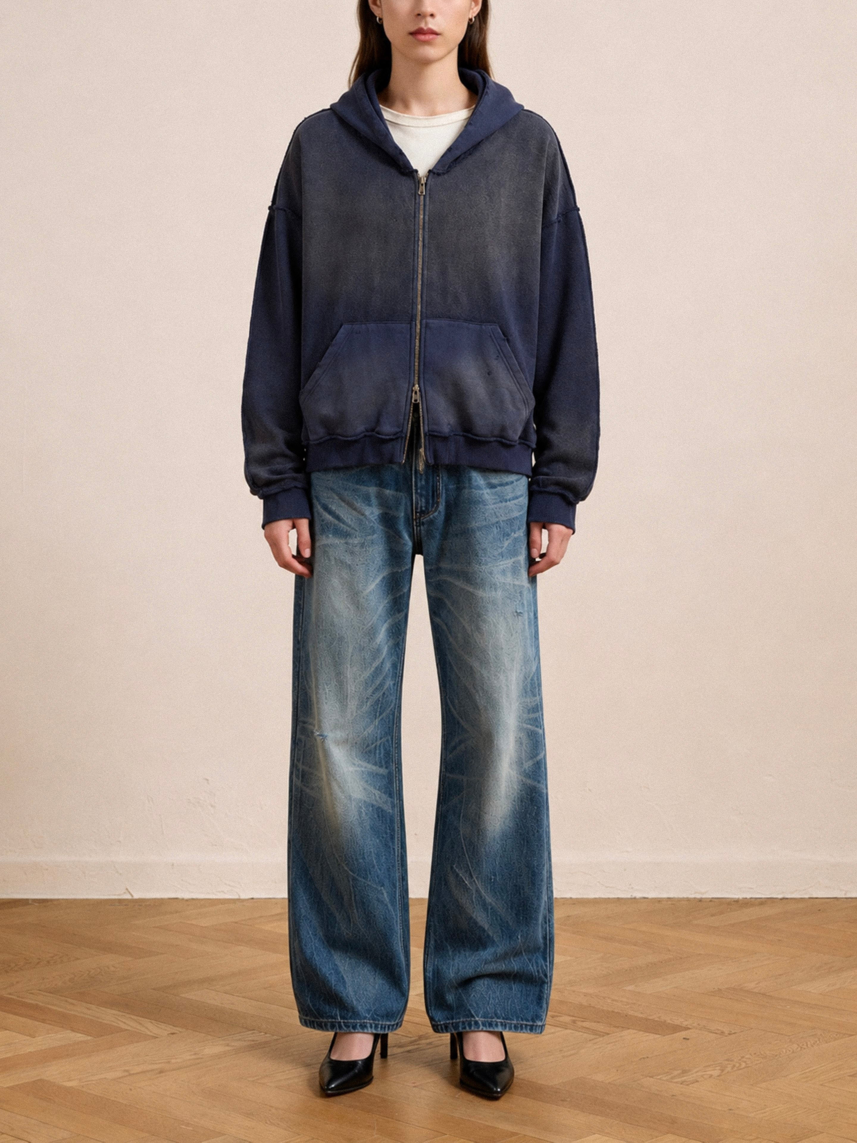 Indoors, a person wears the FOUND Deconstructed Terry Zip Up Hoodie in faded navy, wide-leg blue jeans, and black heels while standing on a wooden floor against a plain wall.