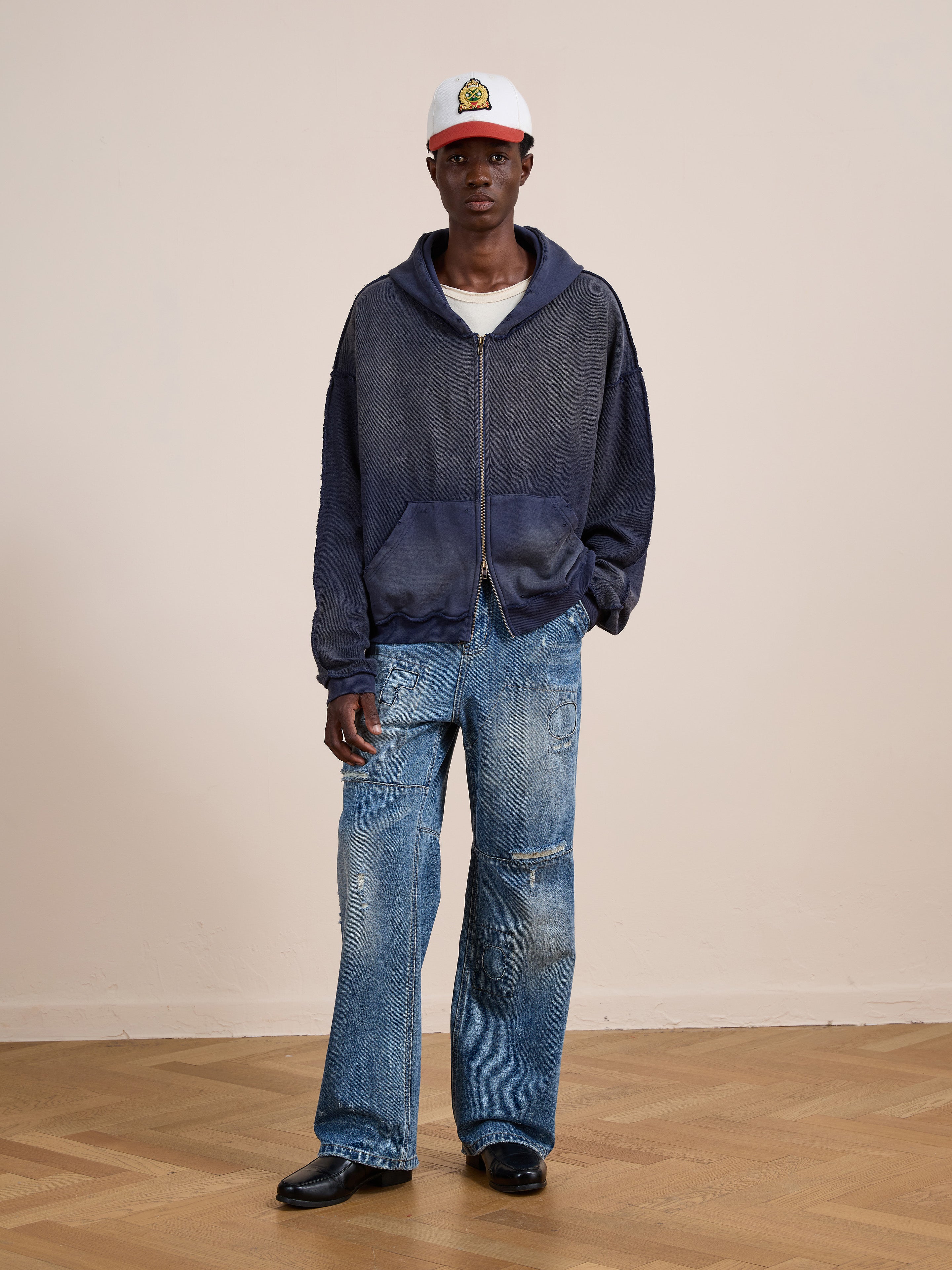 A person stands indoors against a beige wall, wearing a white cap, FOUND Deconstructed Terry Zip Up Hoodie in faded blue, distressed wide-leg jeans, and black shoes on a wooden floor.