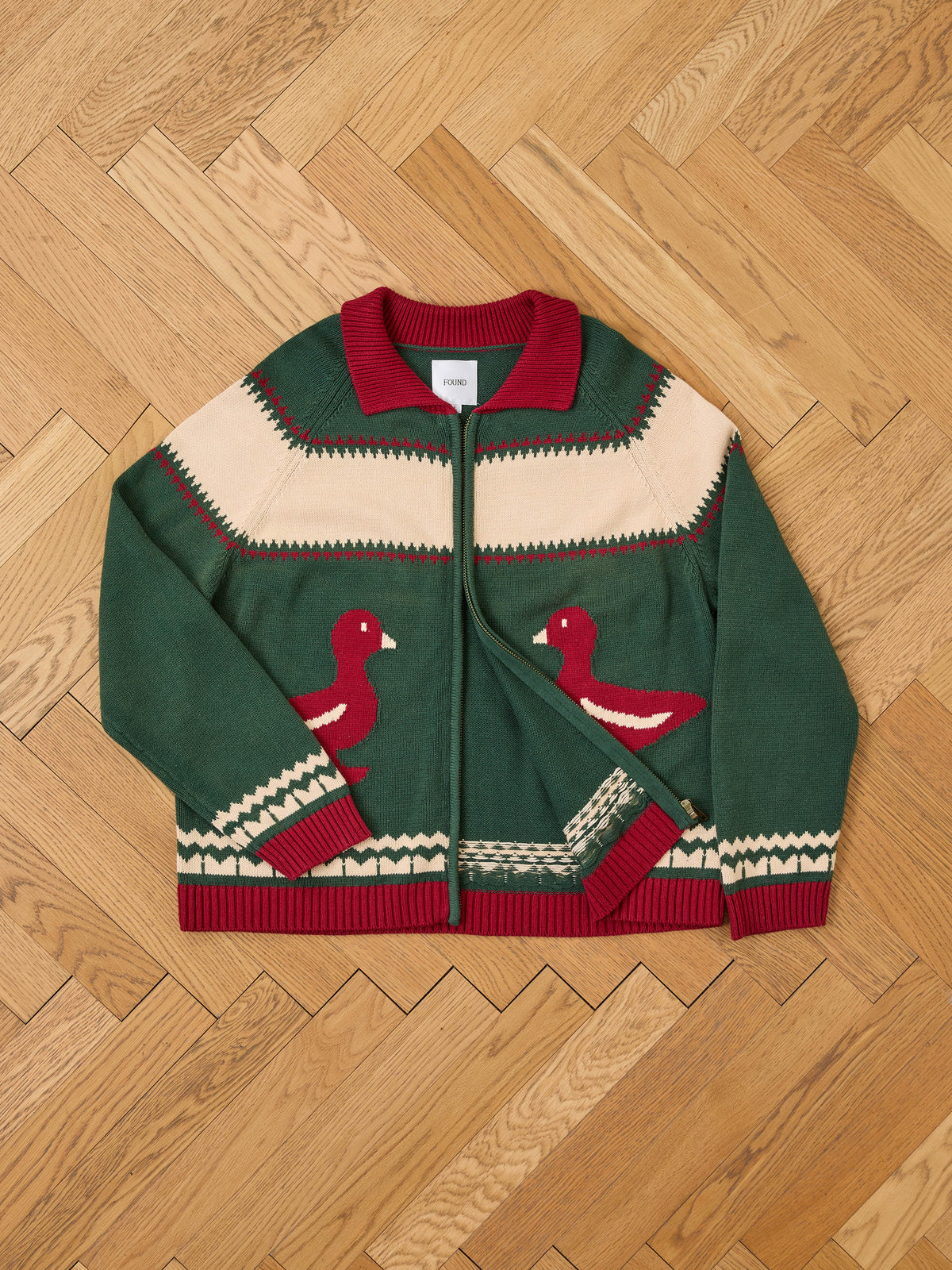 FOUND Isles Sweater: green and cream zip-up with red collar, cuffs, hem, and two red duck motifs on the front, shown flat on a wood herringbone floor.