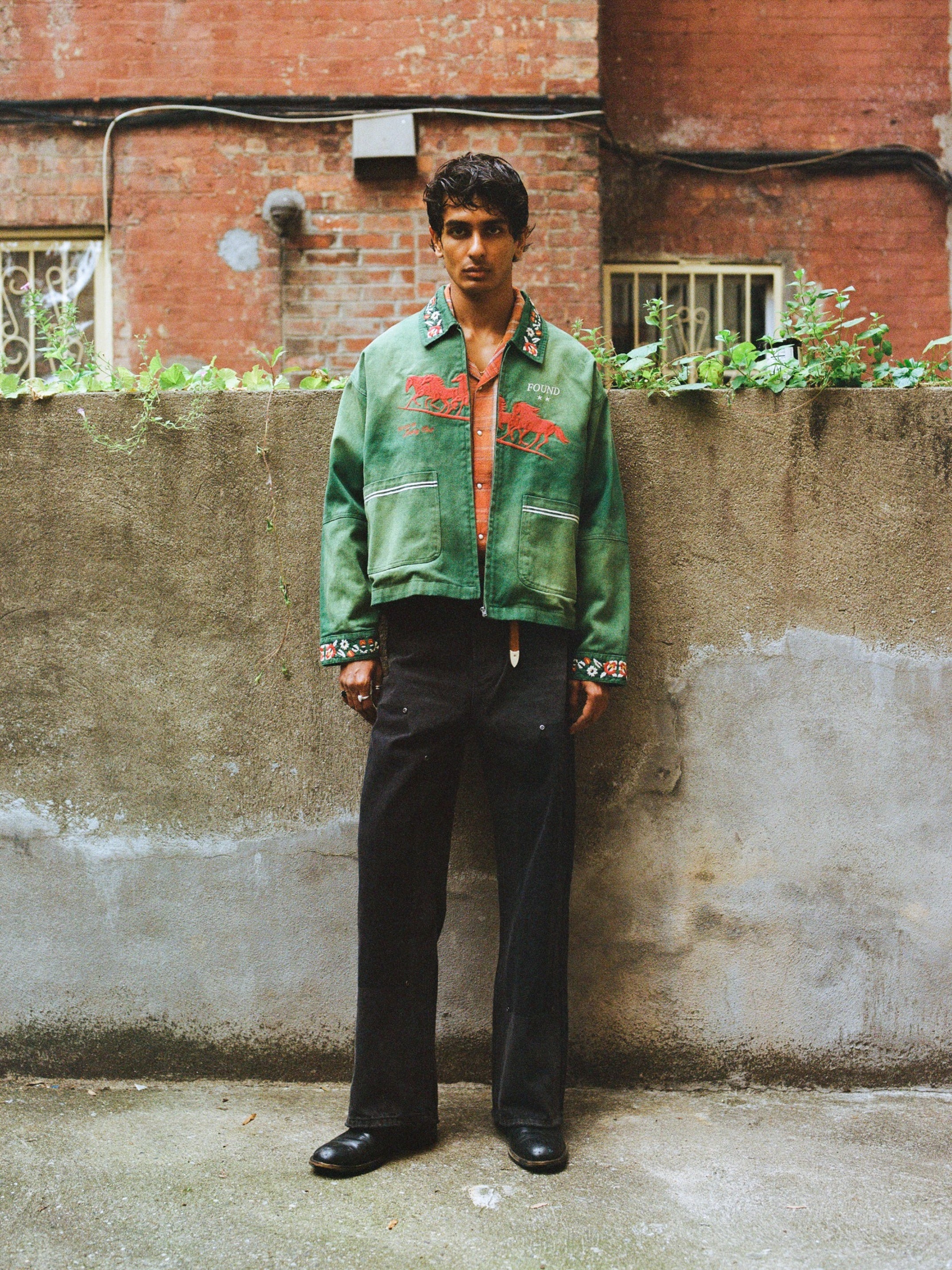 A man stands in front of a concrete wall wearing the Found Jockey Club Embroidered Jacket, an orange shirt, black pants, and black shoes. A red brick building and greenery are visible in the background.