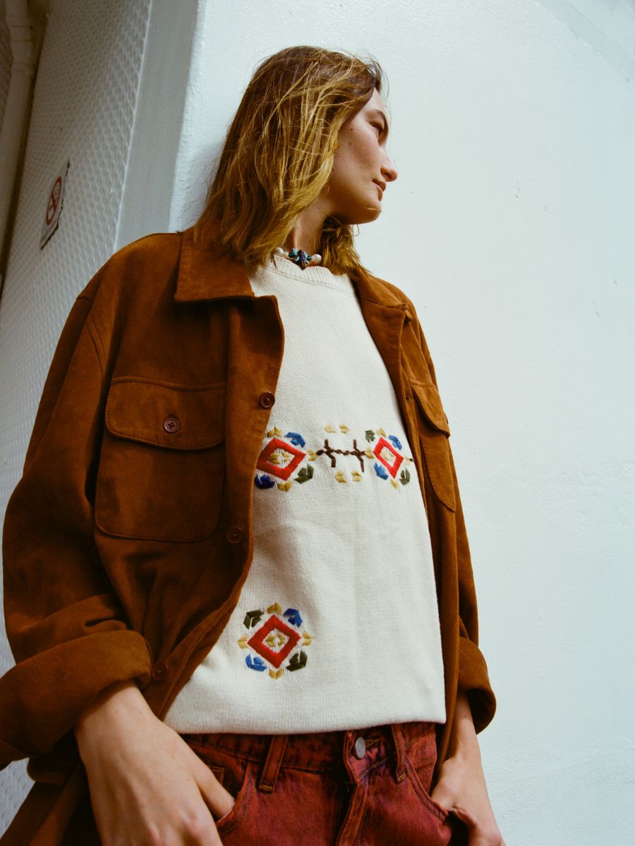 A person wearing FOUND’s Knit Flower Petal Tee under a brown jacket stands against a white wall, glancing to the side and radiating vintage-inspired charm.