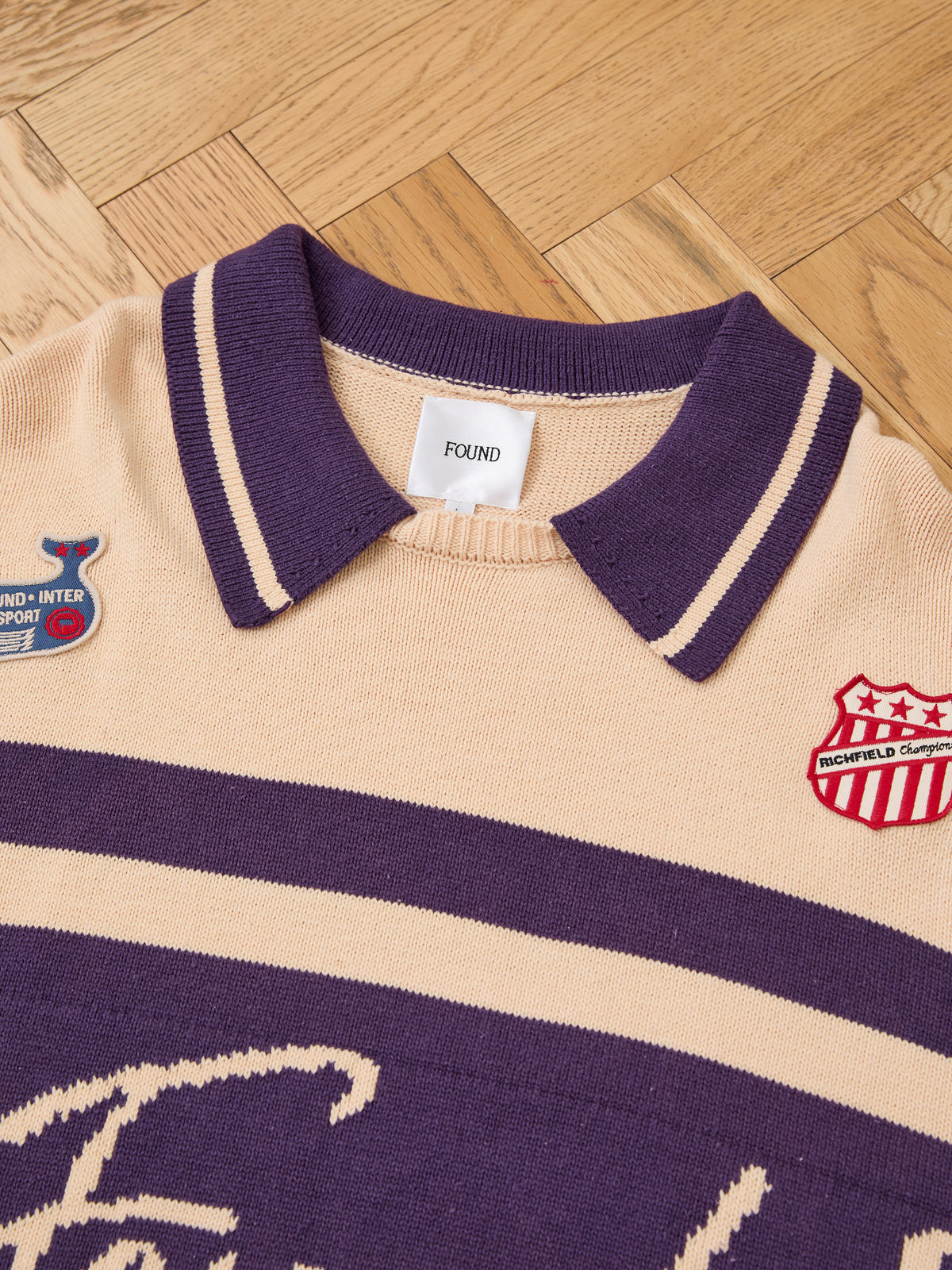A close-up of the FOUND Knit Jersey—beige and purple, vintage club style, with two embroidered patches and a “FOUND” label on a wooden floor.