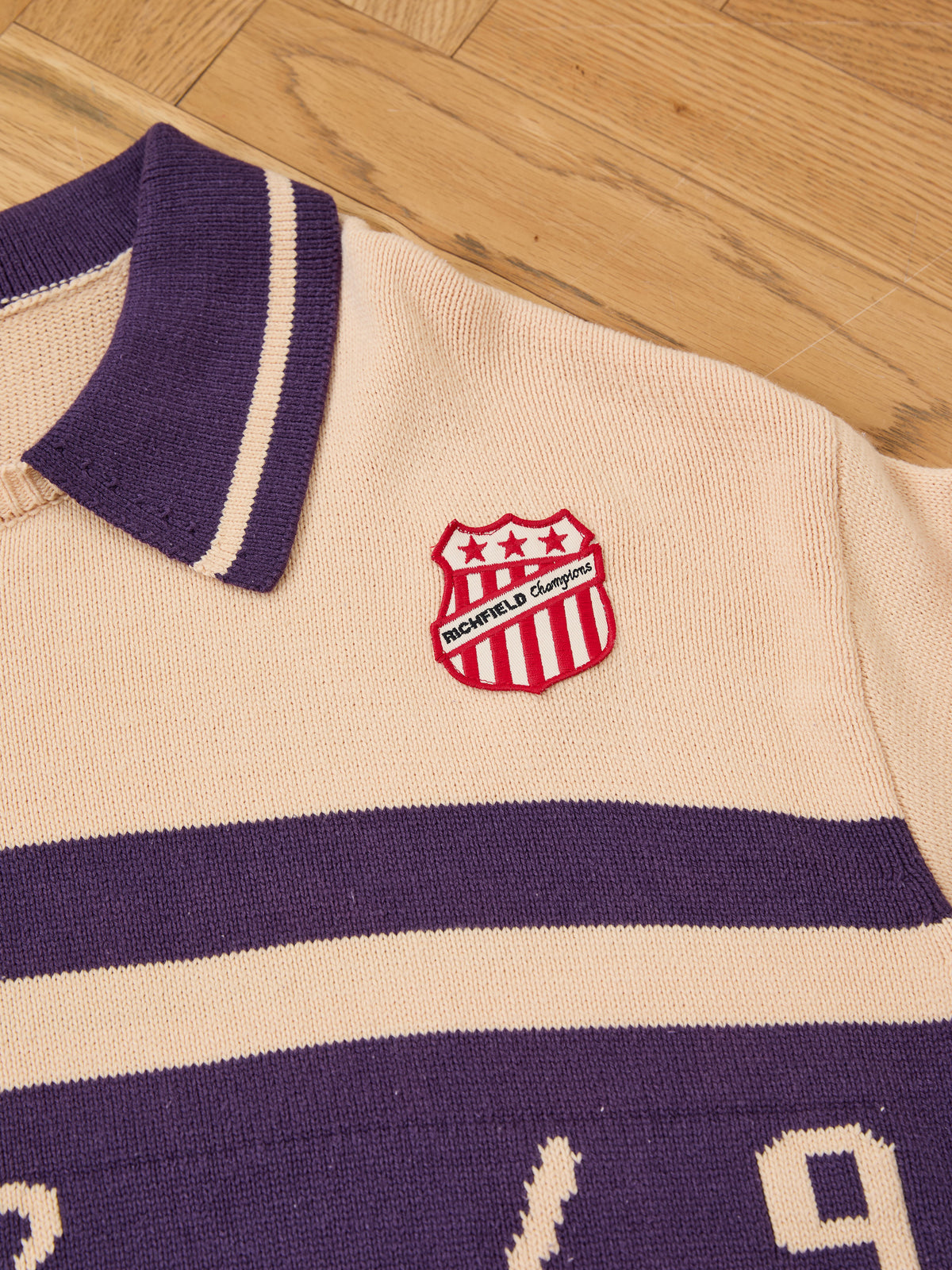 Close-up of a collared beige and purple Knit Jersey by FOUND, featuring a red and white “ROCHEFIELD champions” badge, laid on a wooden floor.