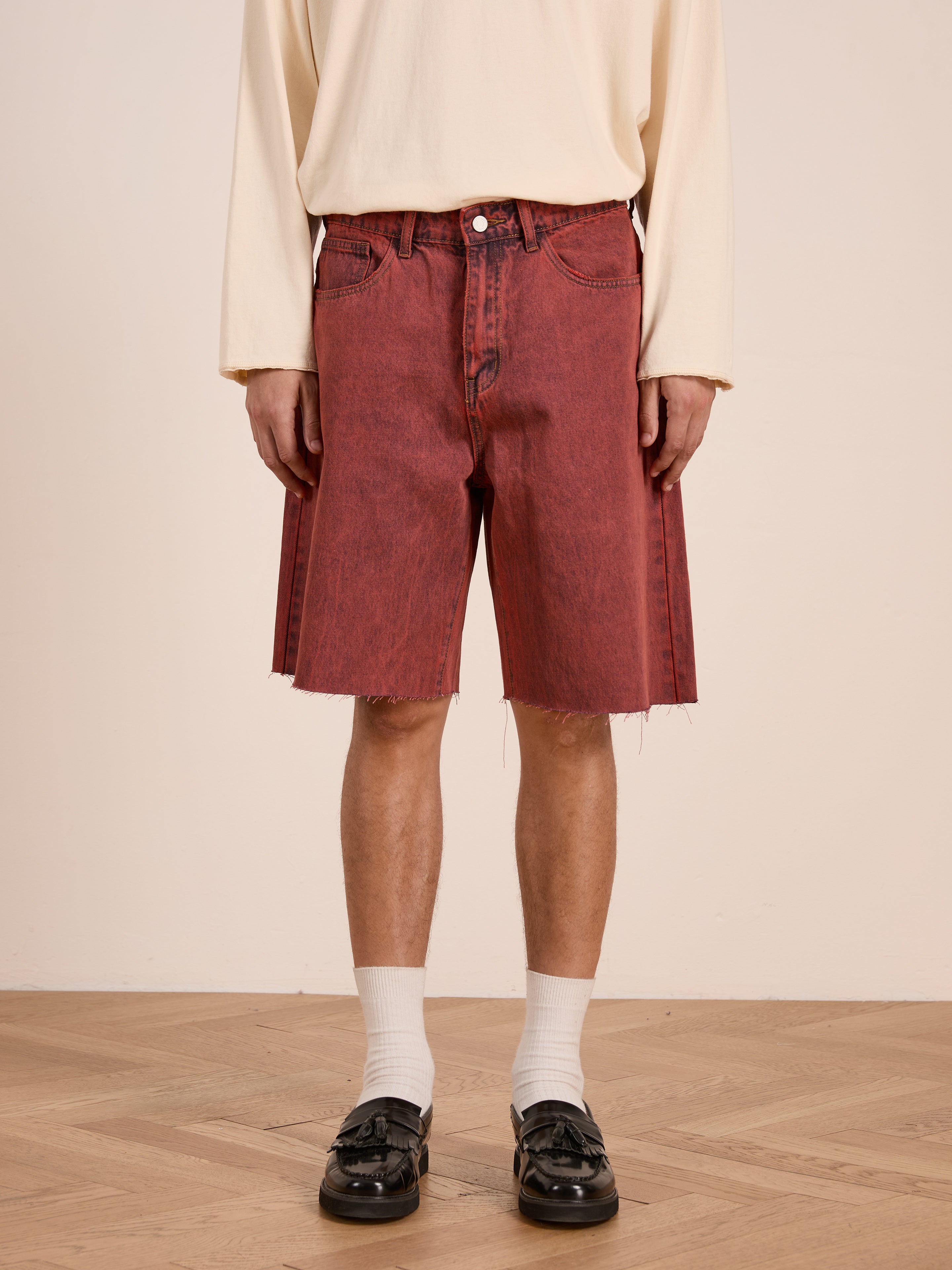 A person wearing a cream long-sleeve shirt, FOUND Lacy Jean Jorts in vintage red wash with frayed hems, white socks, and black loafers stands on a wooden floor.