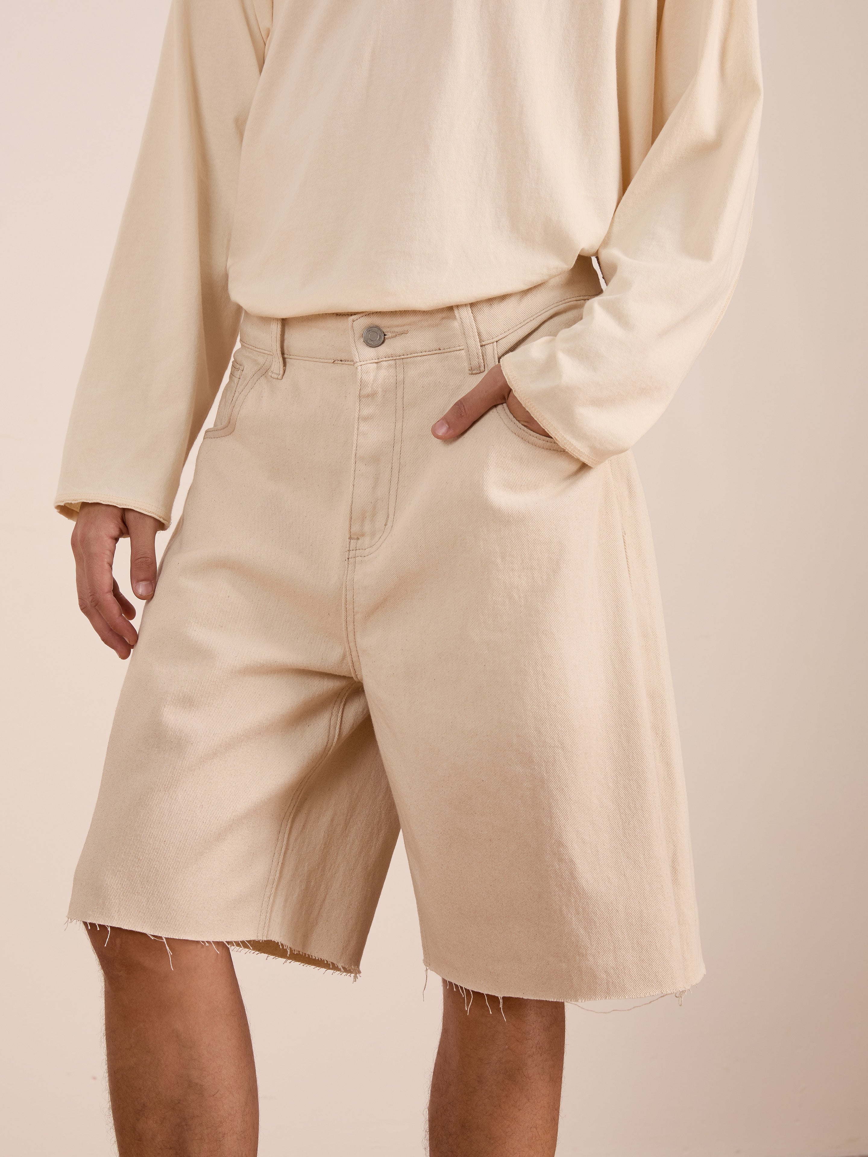 A person models the FOUND Lacy Jean Jorts—distressed denim shorts with a raw hem—paired with a long-sleeve beige shirt, standing with one hand in their pocket.