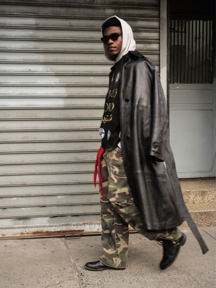 A person wearing the FOUND Leather Trench Coat, camo pants, black shoes, and sunglasses walks on a city sidewalk in front of a metal shutter.