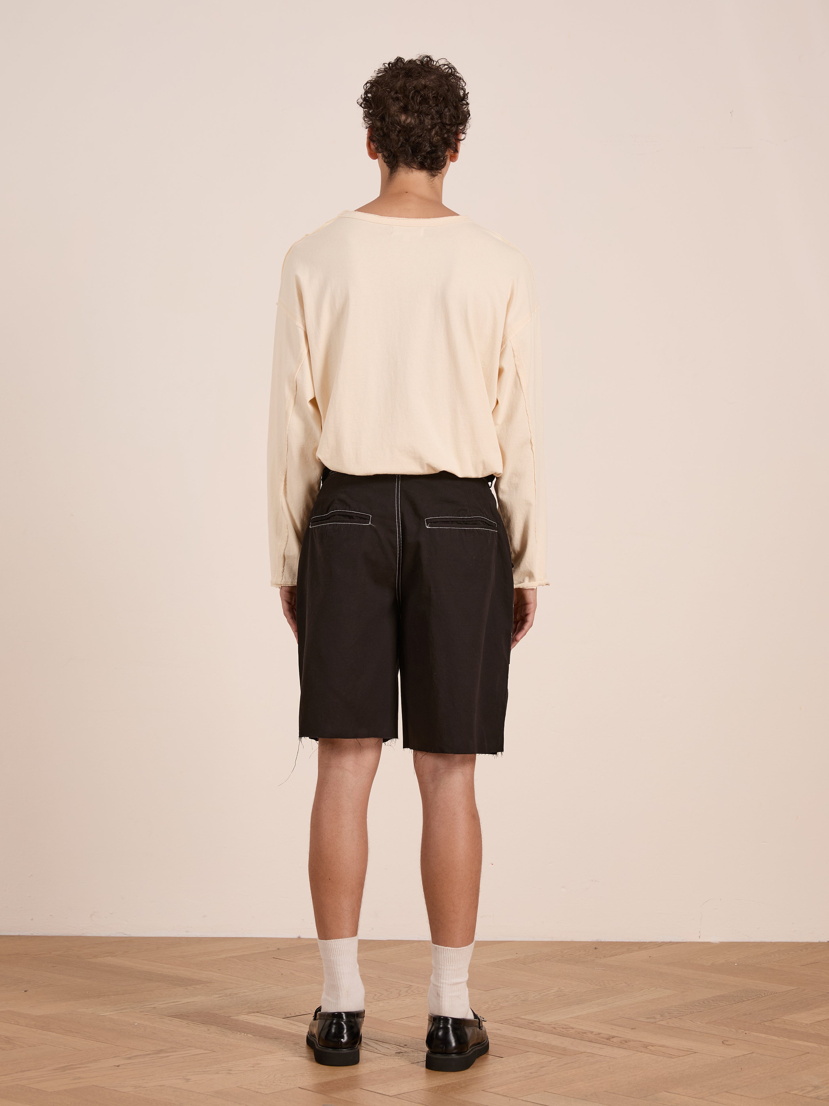 A person stands facing away from the camera, wearing a cream long-sleeve shirt and FOUND Lucca Black Contrast Pocket Trouser Shorts made from cotton twill, paired with white socks and black shoes on a wooden floor for a vintage-inspired look.