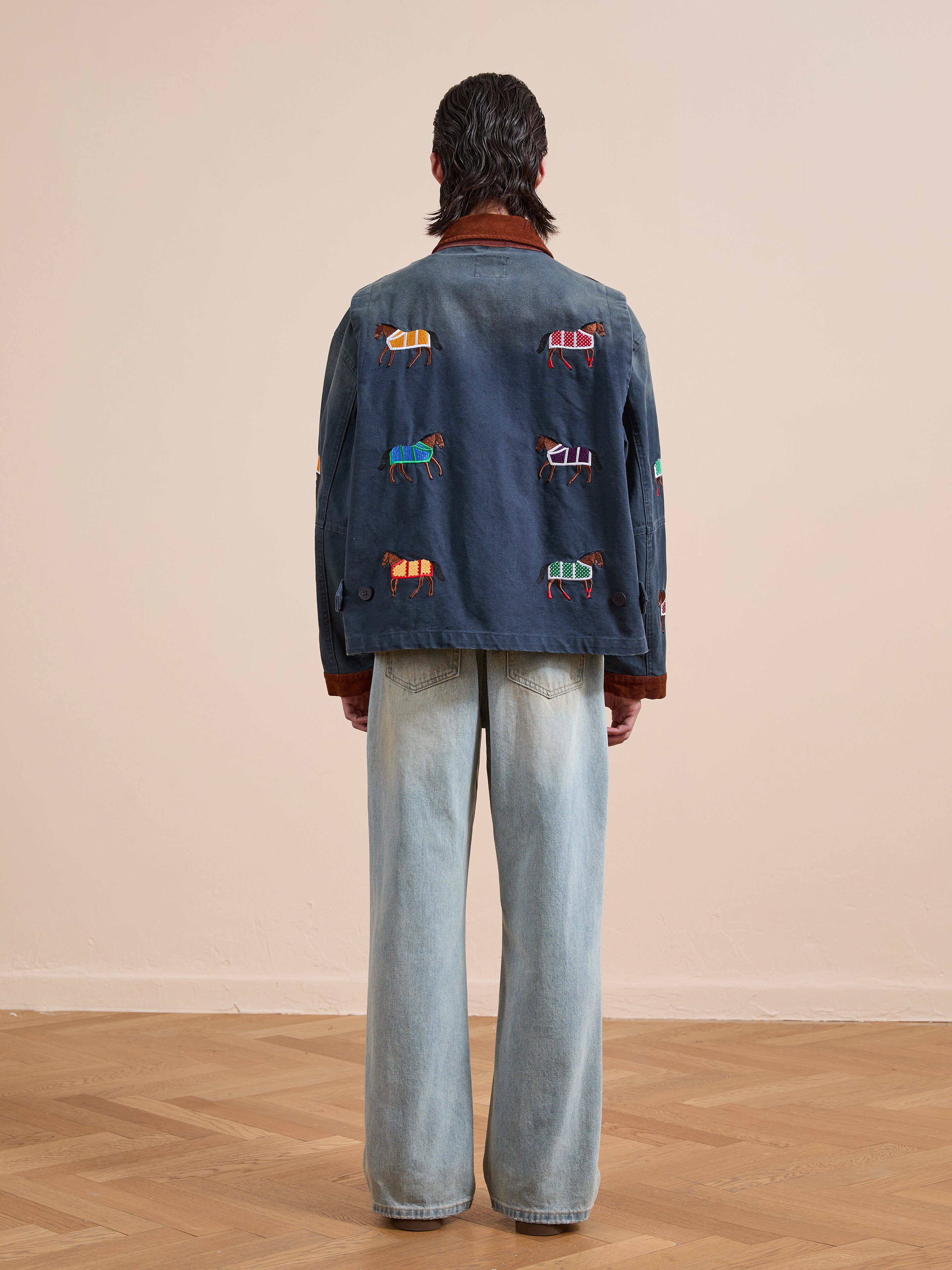 A person stands indoors with a plain light wall and wood floor, wearing Found’s Lariat Horse Embroidered Work Jacket in navy with colorful horse embroidery on the back, paired with light blue wide-leg jeans.