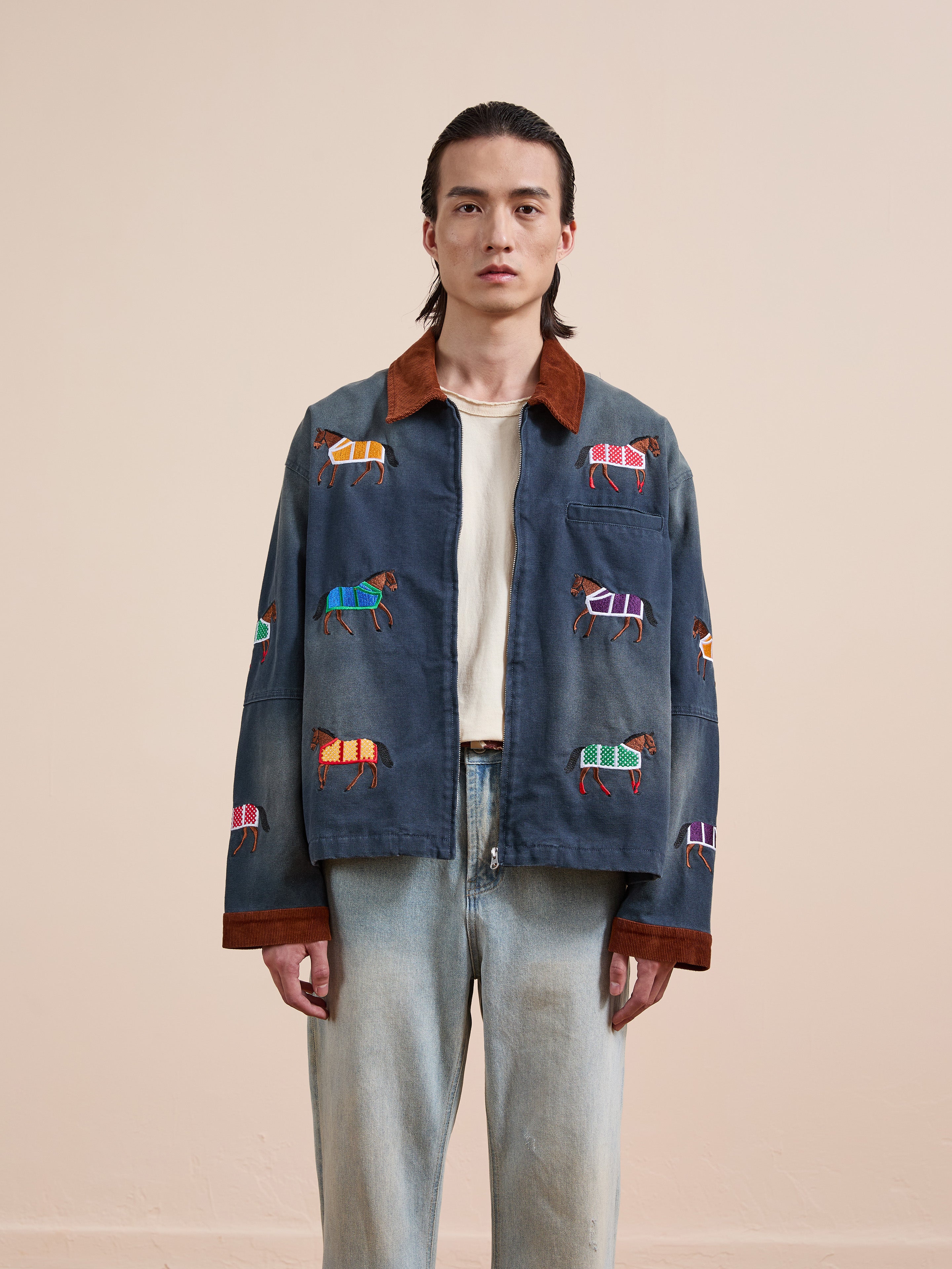 A person stands facing forward against a beige background, wearing the Found Lariat Horse Embroidered Work Jacket— a unisex navy piece with colorful animal patterns, horse embroidery, brown collar—paired with a white shirt and light blue jeans.