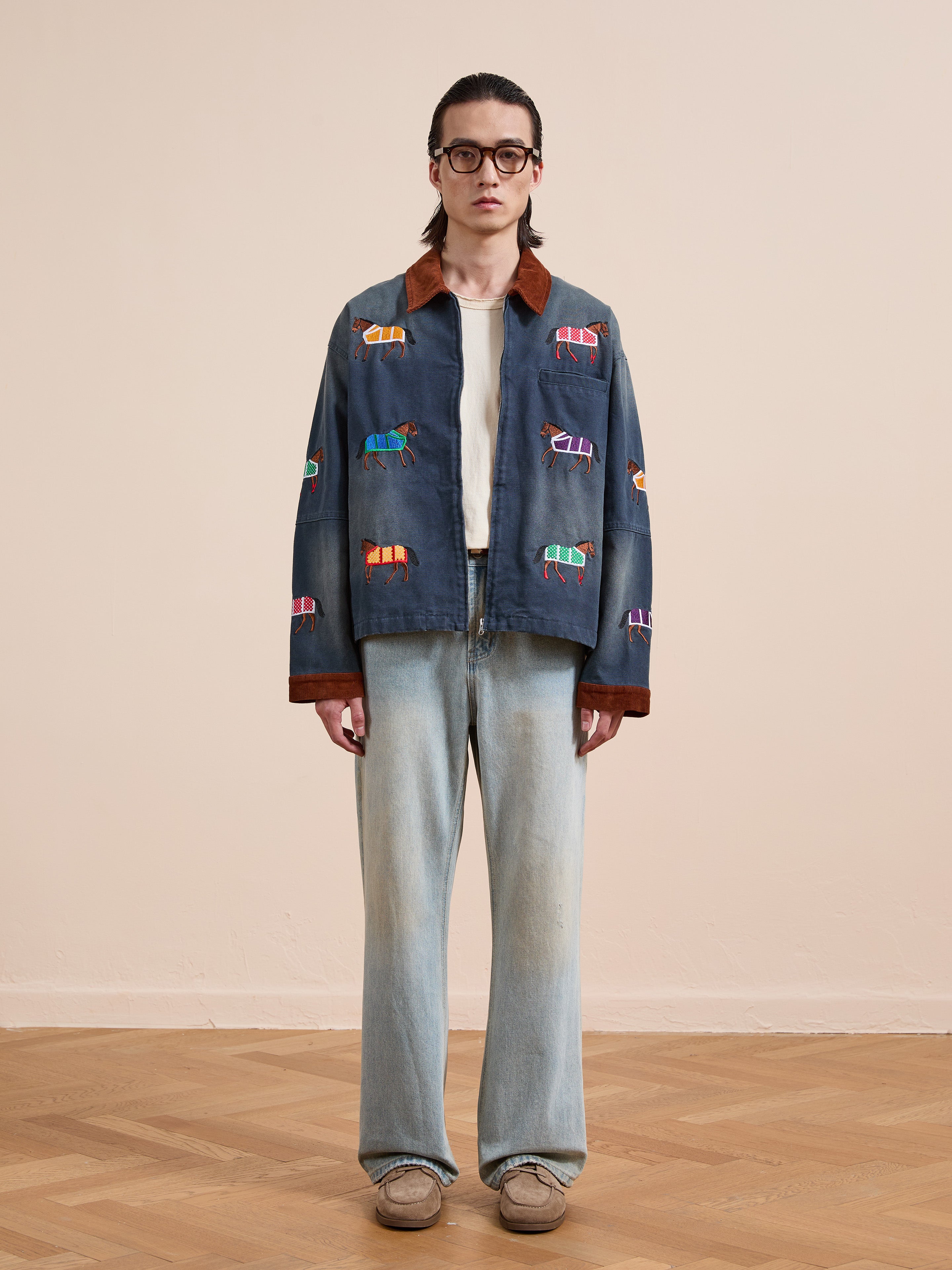 A person stands indoors wearing glasses, a white shirt, light-wash jeans, beige shoes, and the Found Lariat Horse Embroidered Work Jacket on a wooden floor.