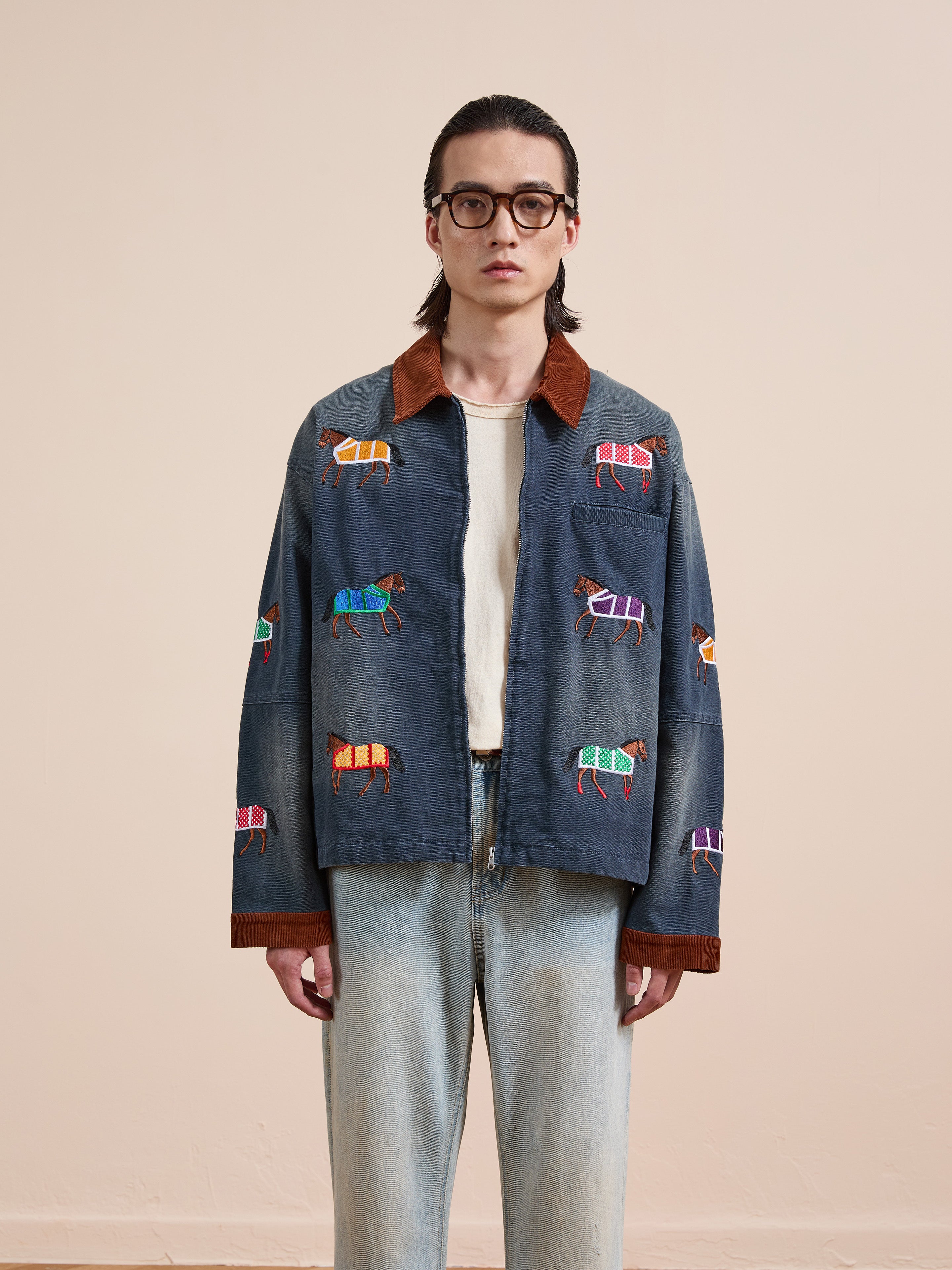 Someone wearing glasses, the Found Lariat Horse Embroidered Work Jacket in navy, a white shirt, and light blue jeans stands against a plain beige background.