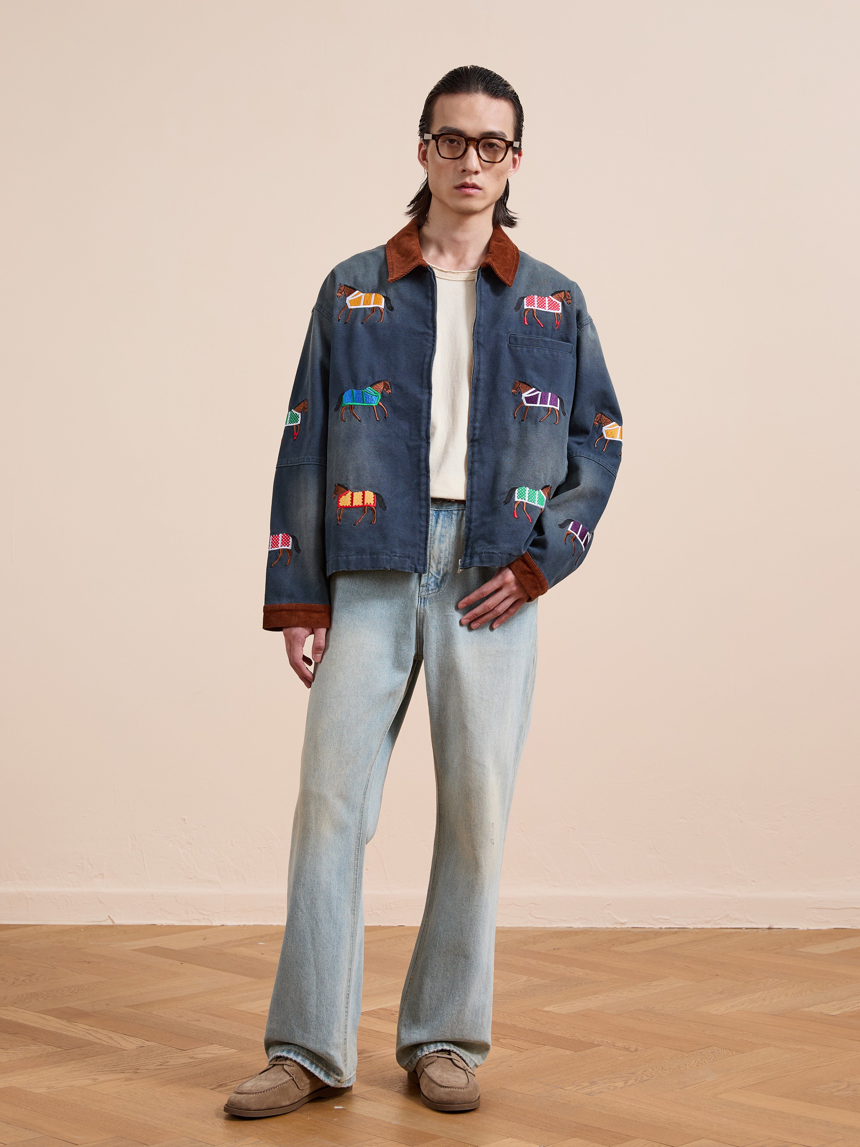 A person stands indoors on a wooden floor wearing glasses, the Found Lariat Horse Embroidered Work Jacket, a white shirt, light blue jeans, and beige shoes.