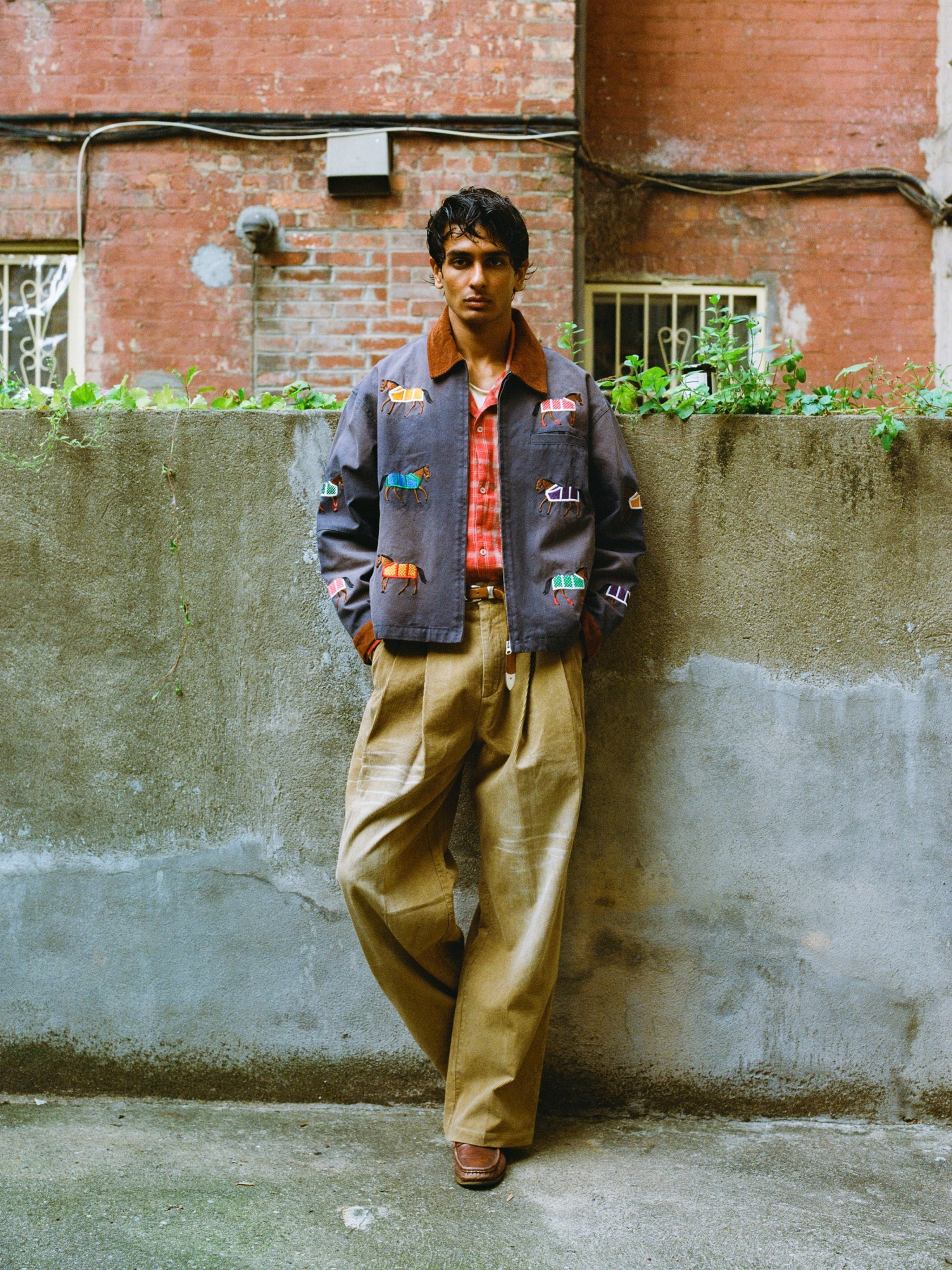 A person stands by a concrete wall in an urban outdoor setting, wearing Found's Lariat Horse Embroidered Work Jacket, a red plaid shirt, and wide khaki pants.