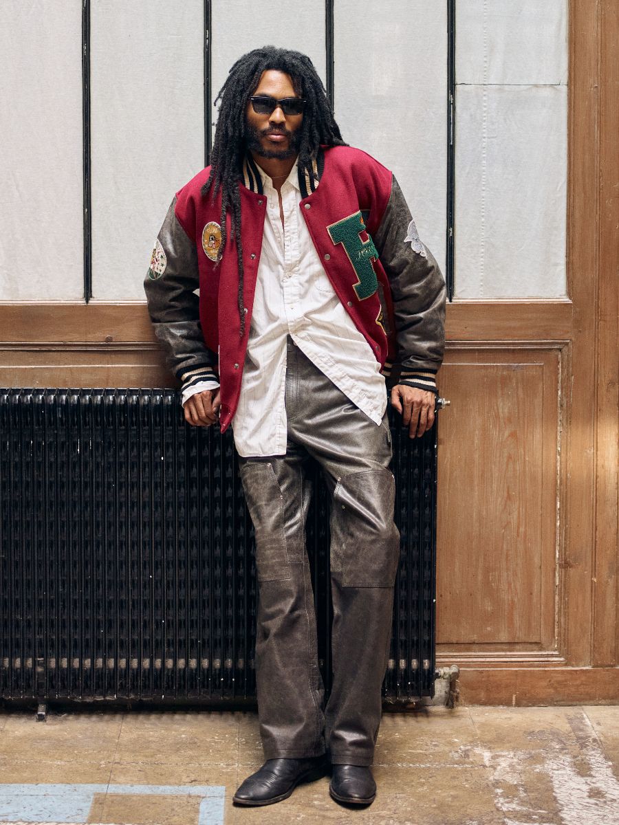 A person in sunglasses and dreadlocks leans against a wall, wearing a red varsity jacket, white shirt, and unisex relaxed fit distressed genuine leather carpenter pants by FOUND. They strike a confident pose in boots on a wooden floor, showcasing effortless style.