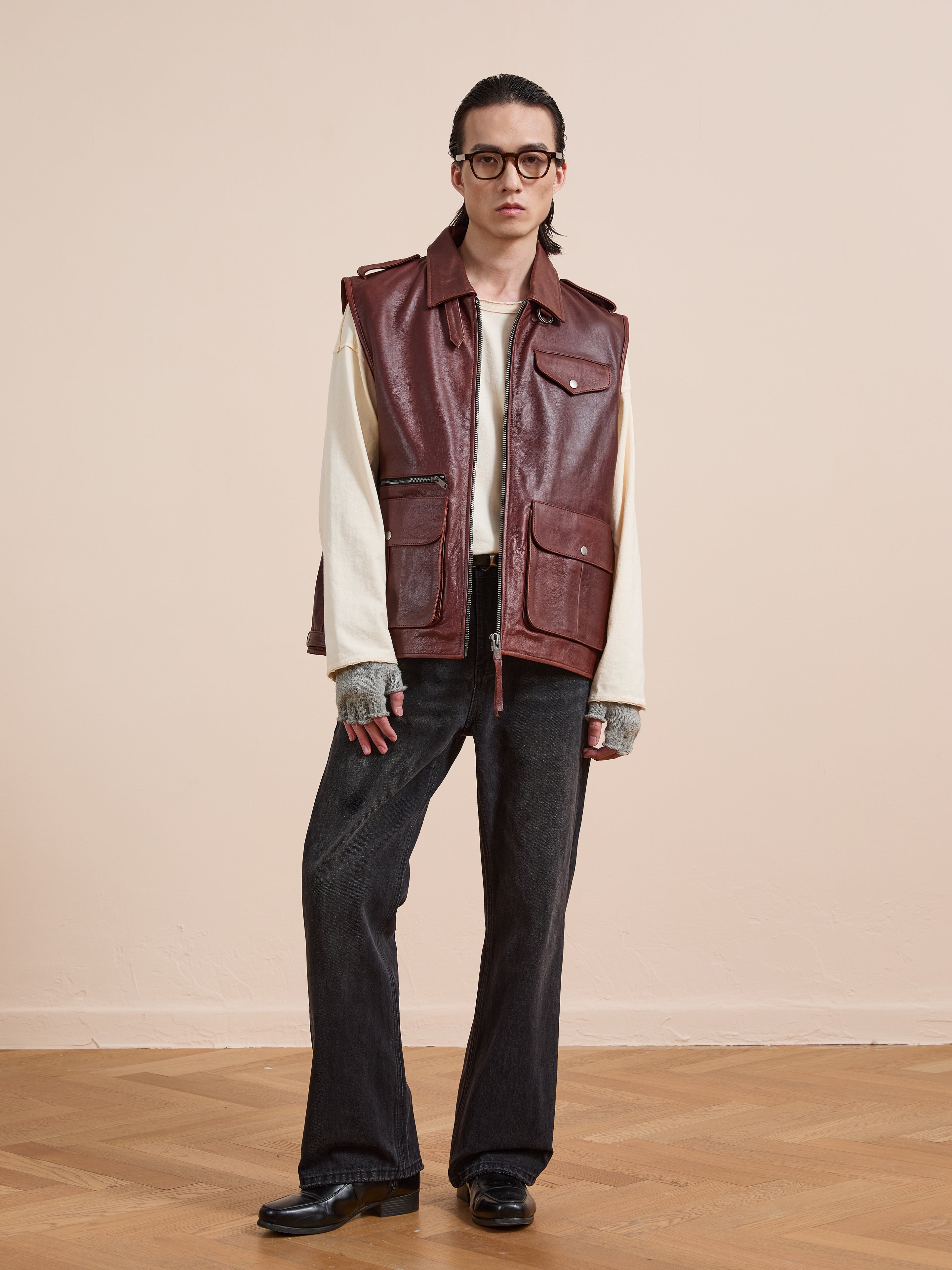 A person stands indoors wearing Found’s distressed brown Leather Pocket Vest with utility pockets over a white long-sleeve shirt, loose dark jeans, gray fingerless gloves, black shoes, glasses, and slicked-back hair.
