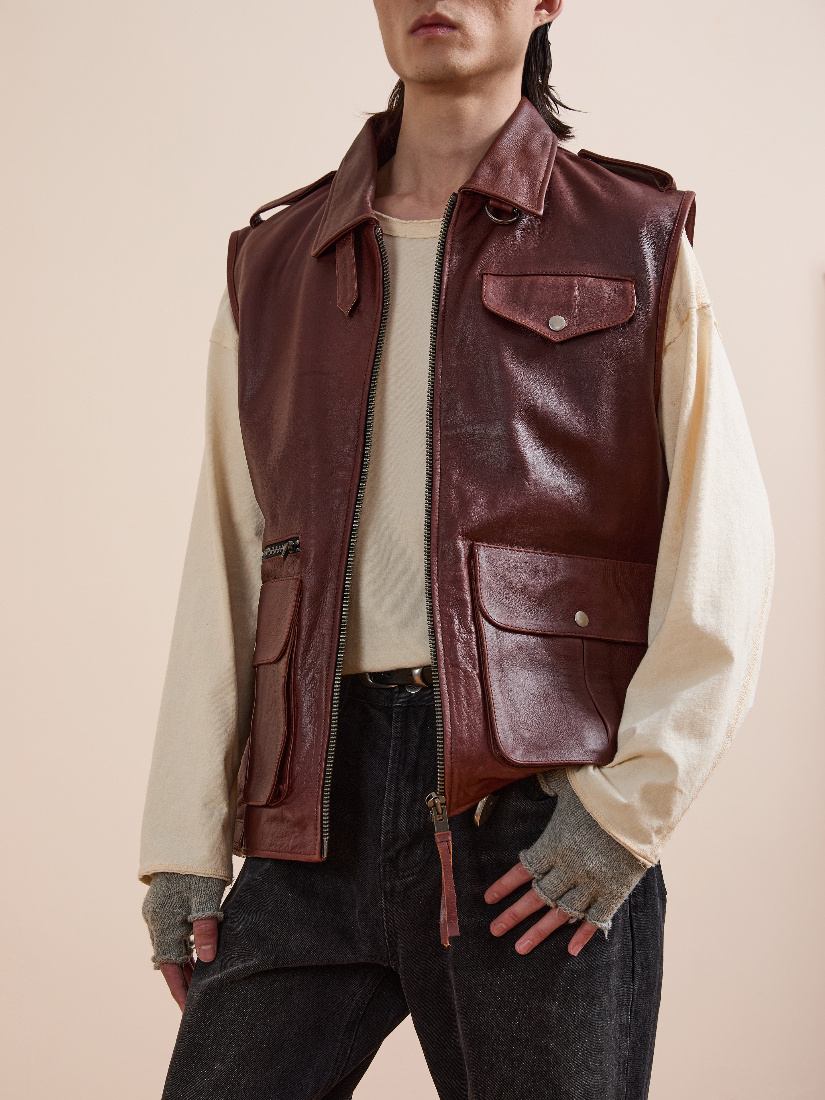 A person wears Found's Leather Pocket Vest in distressed brown with utility pockets over a beige long-sleeve shirt, fingerless gloves, and black jeans, standing against a plain background.