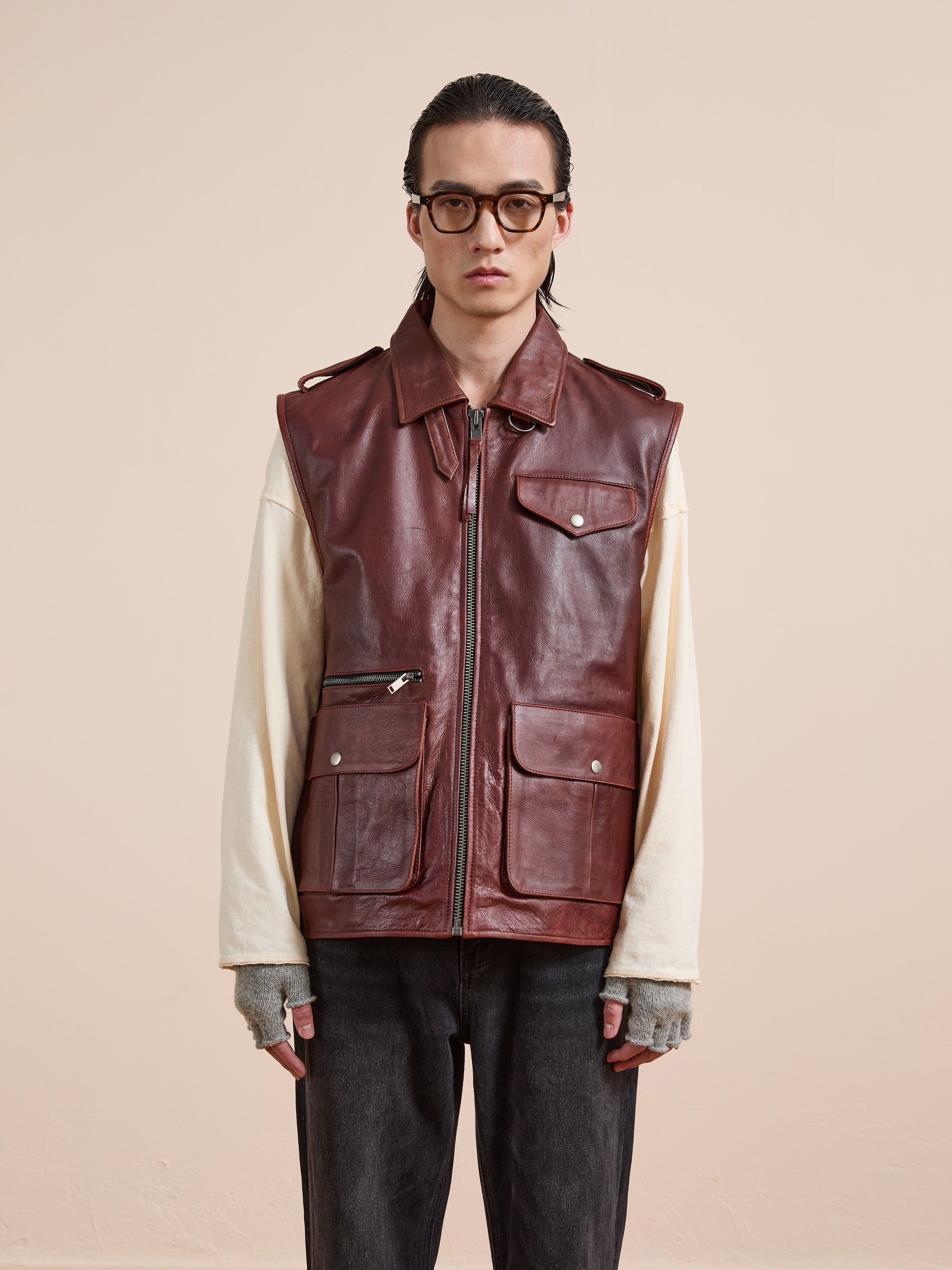 A person wearing glasses, the Found Leather Pocket Vest with utility pockets over a beige long-sleeve shirt, gray fingerless gloves, and dark pants stands against a plain beige background.